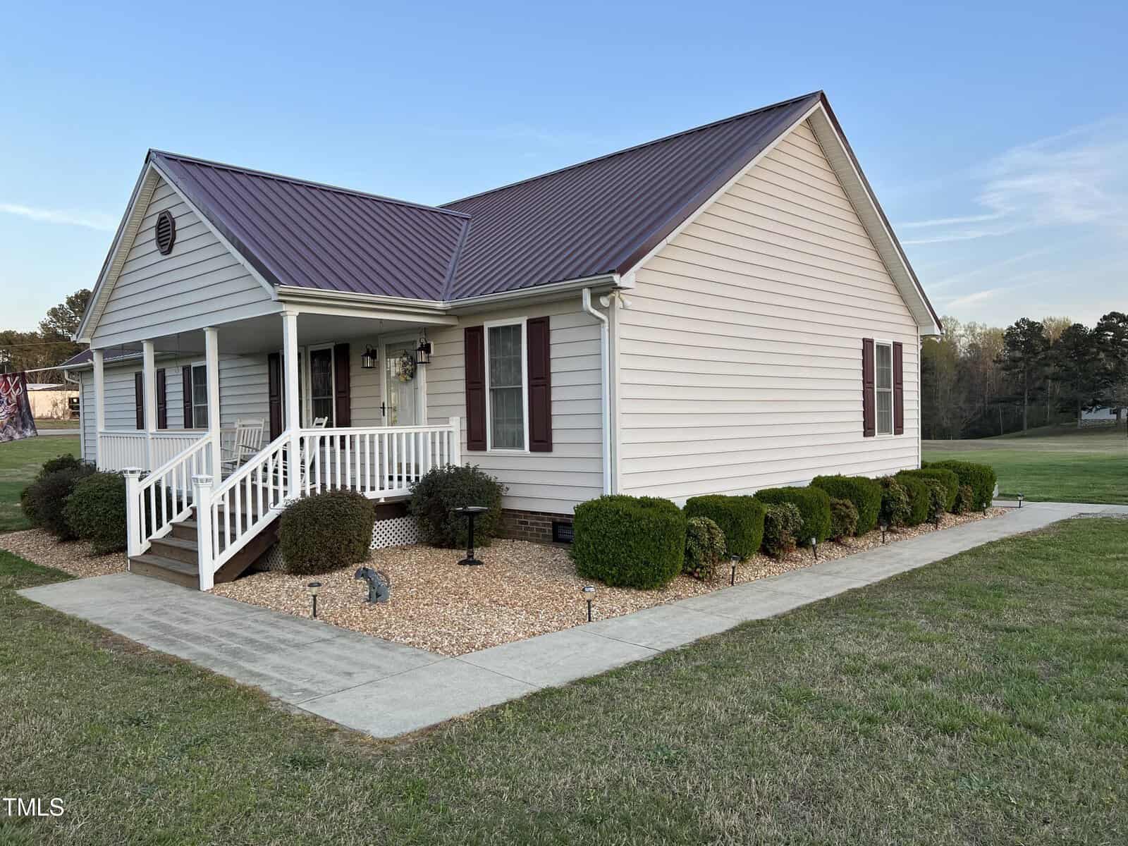 A one-story beige house with a white porch and railing, surrounded by small, neatly trimmed bushes and a gravel front yard, set against a backdrop of a clear sky at twilight.