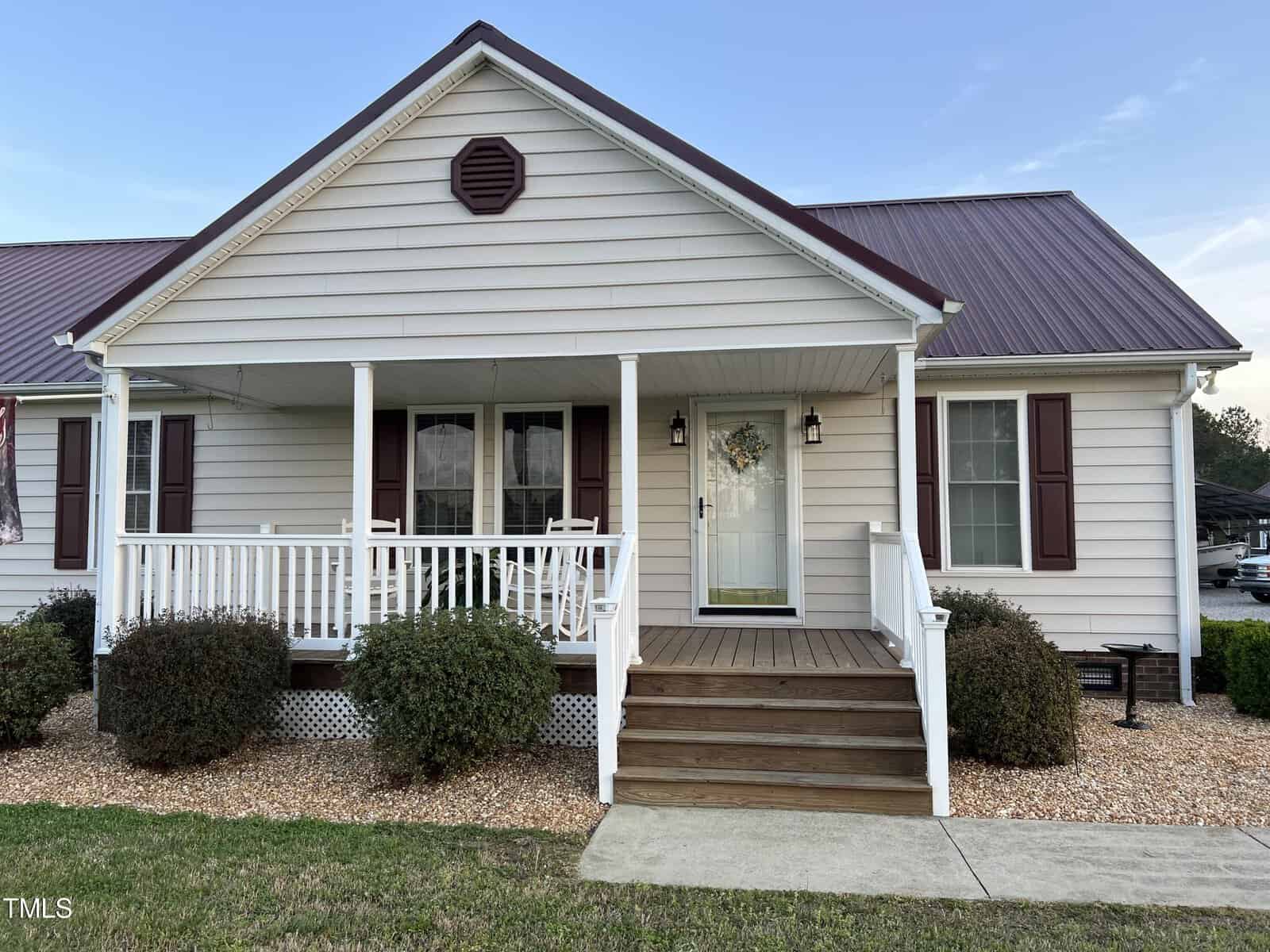 A charming single-story house with beige siding and dark red shutters, featuring a covered front porch with white railings and a welcoming wreath on the door.