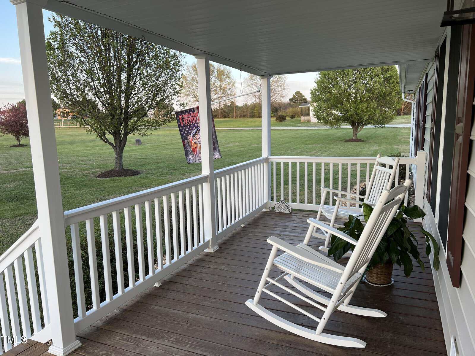 Covered porch with white railings and two rocking chairs facing a lush green lawn with trees and clear skies, giving a serene suburban feel.