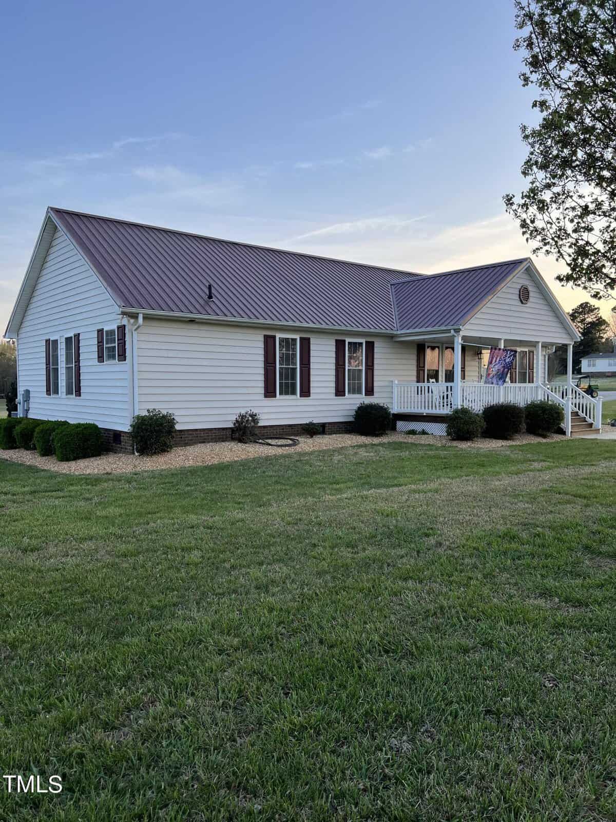 A one-story suburban home with a gabled metal roof, white siding, and a front porch. the house is surrounded by a manicured lawn and set against a clear sky at dusk.