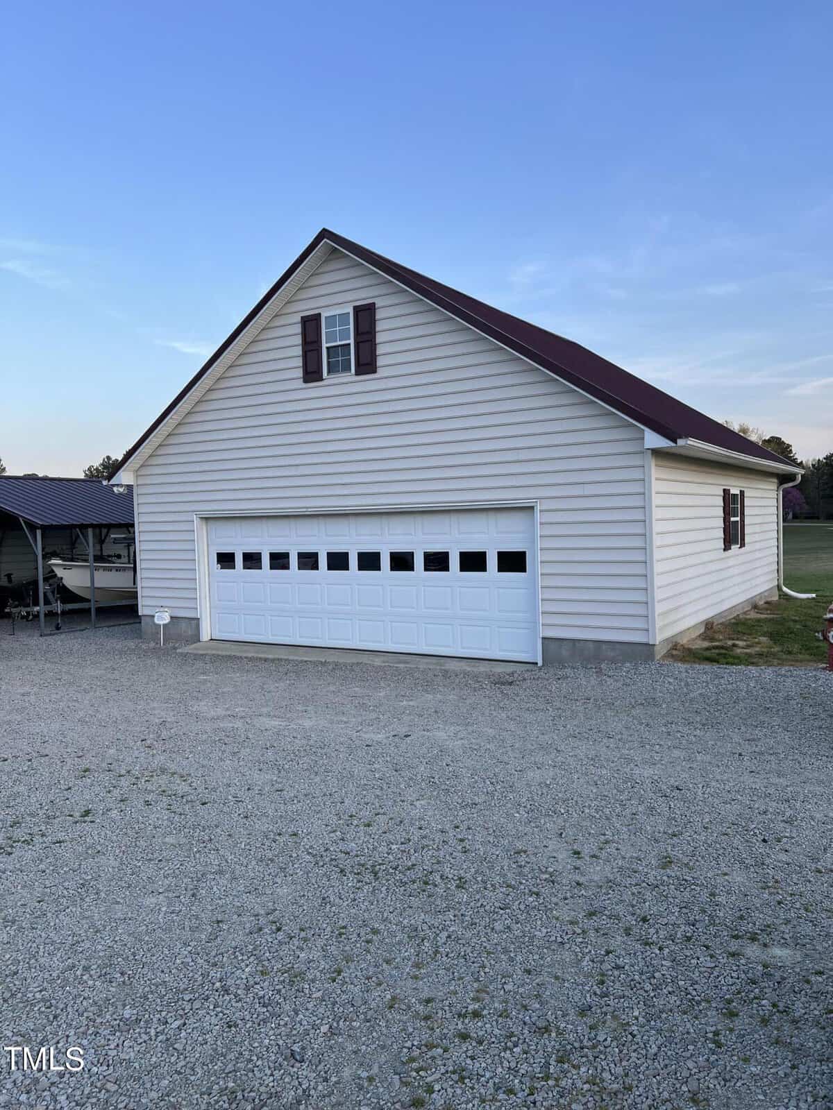 A large detached garage with a white exterior and a gray roof, featuring a two-car garage door. it's surrounded by a gravel driveway and there are trees in the background under a clear sky.