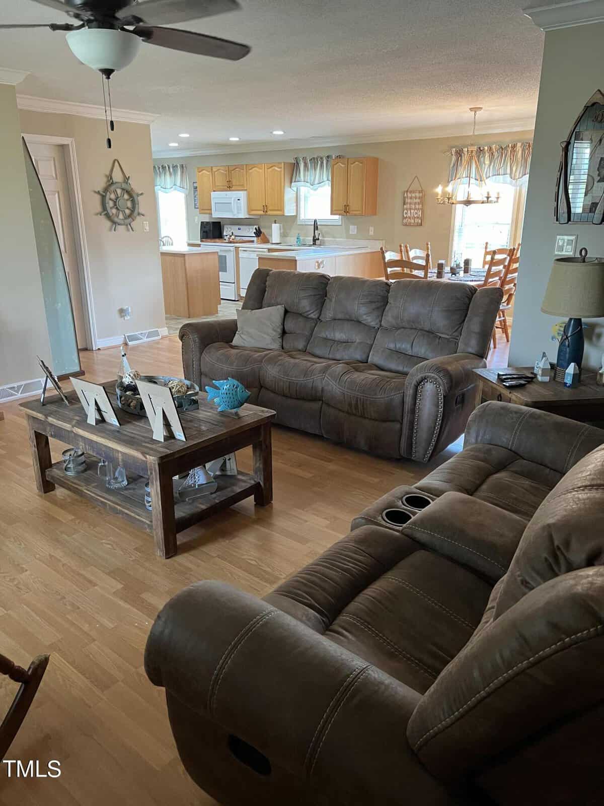 A cozy living room featuring two plush brown sofas, a wooden coffee table, and an open view into a kitchen with light wood cabinetry and a dining area. decor includes a ceiling fan and minimalistic ornaments.