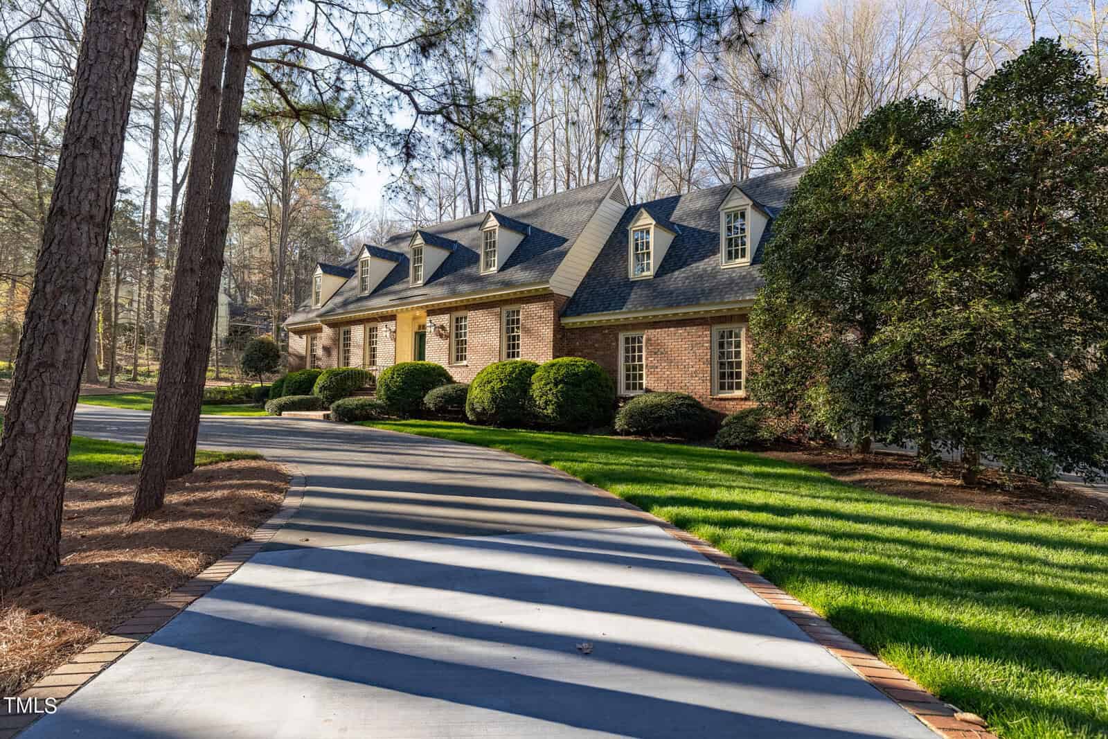 A large brick house with dormer windows surrounded by tall pine trees, featuring a curving sidewalk casting long shadows in the sunlight.