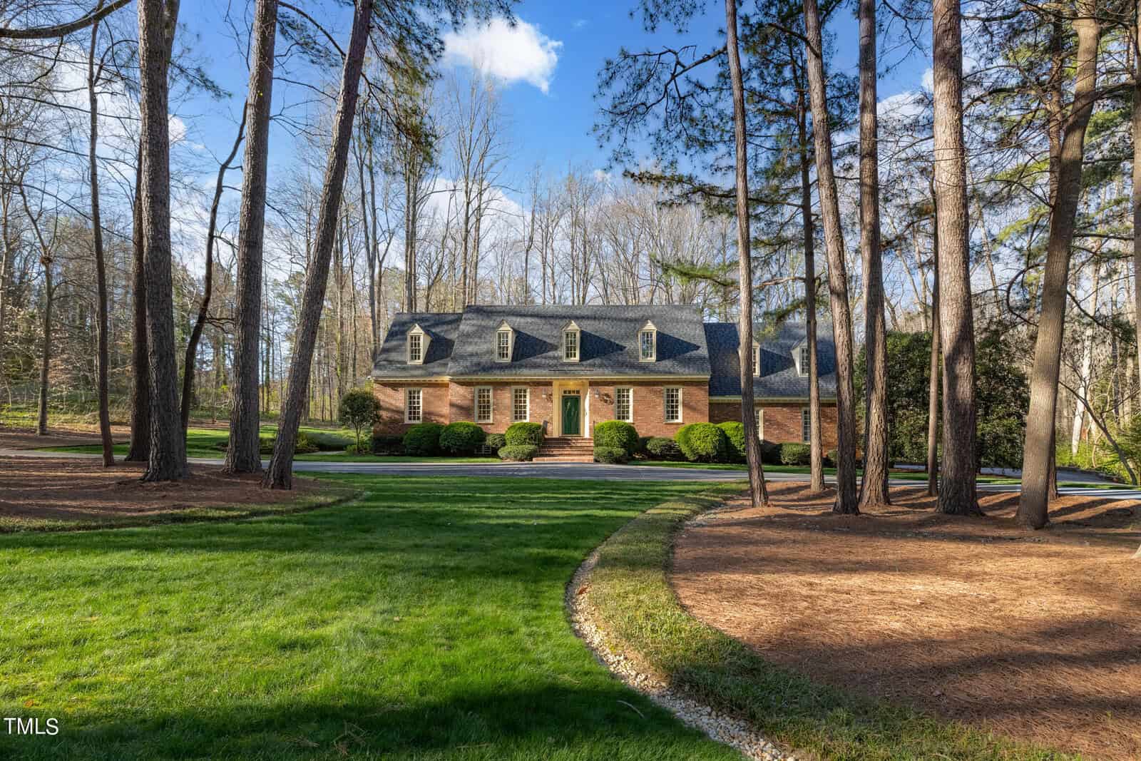 A traditional brick two-story house at 233 Bellwood Drive, Henderson NC, surrounded by tall pine trees and well-maintained lawns with a curved driveway on a sunny day.