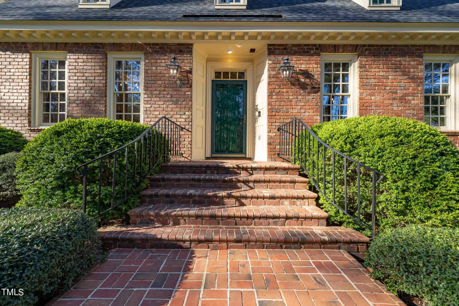 Brick house front entrance with a green door, flanked by manicured bushes and a curving brick staircase, under a sunny sky.
