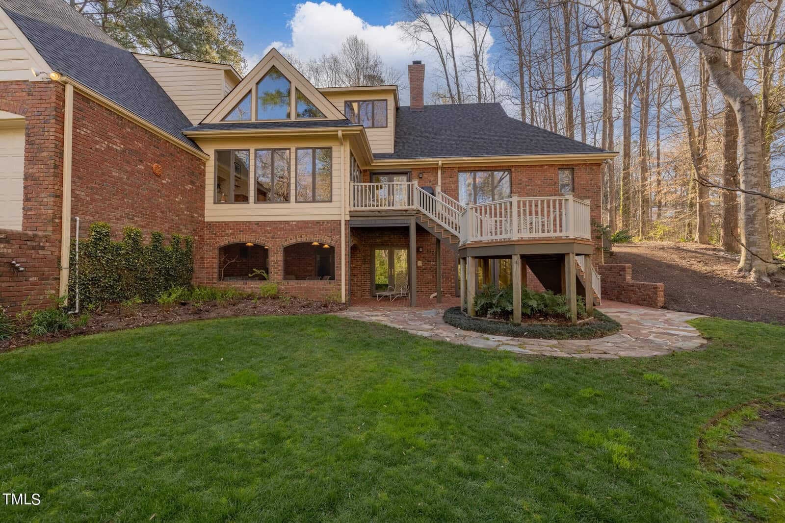 A large brick house with multiple peaked roofs and extensive decking, situated on a manicured lawn with wooded surroundings under a clear sky.
