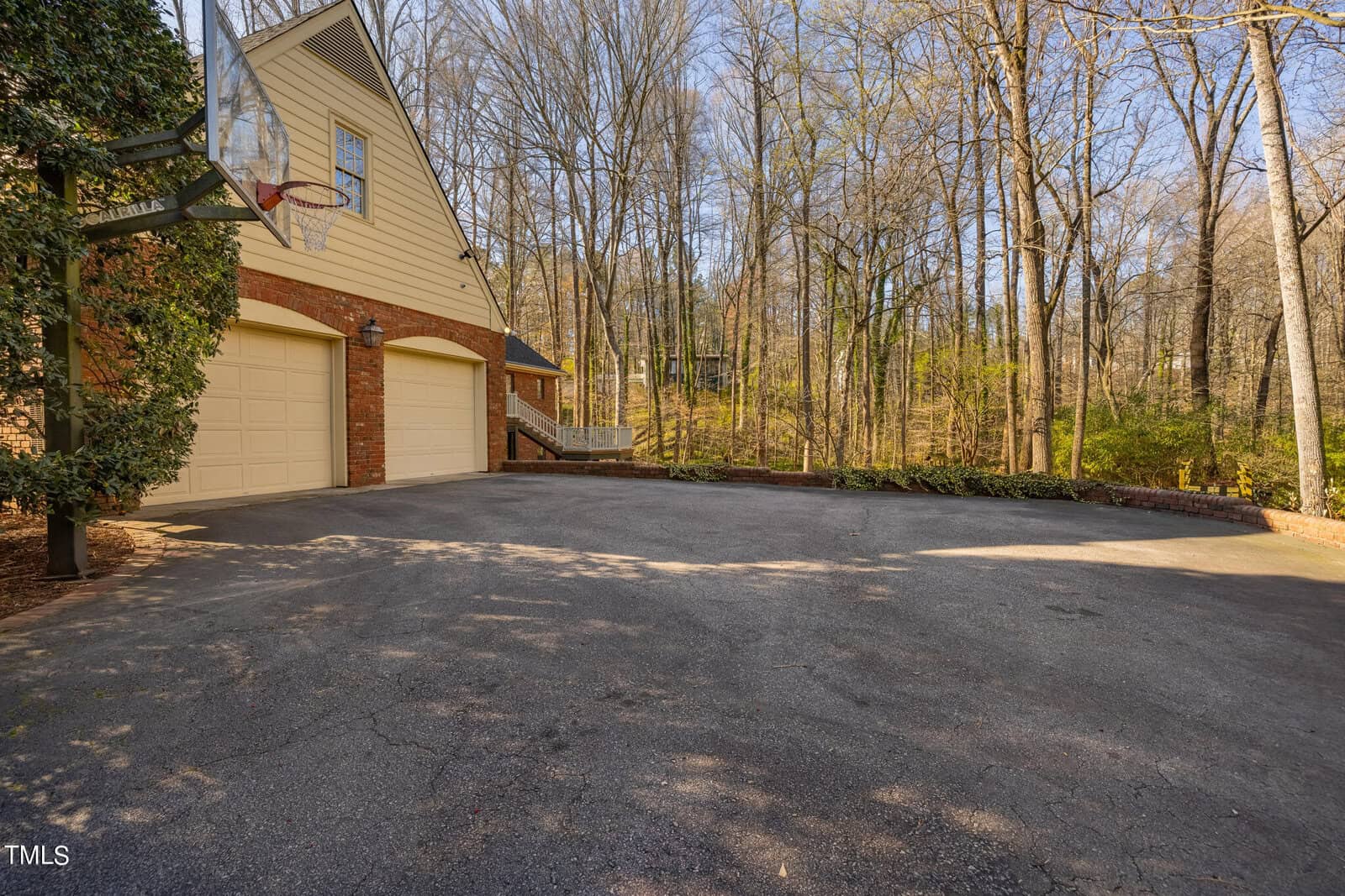 A spacious driveway leads to a two-car garage attached to a house surrounded by leafy trees, with a basketball hoop mounted above the garage door.