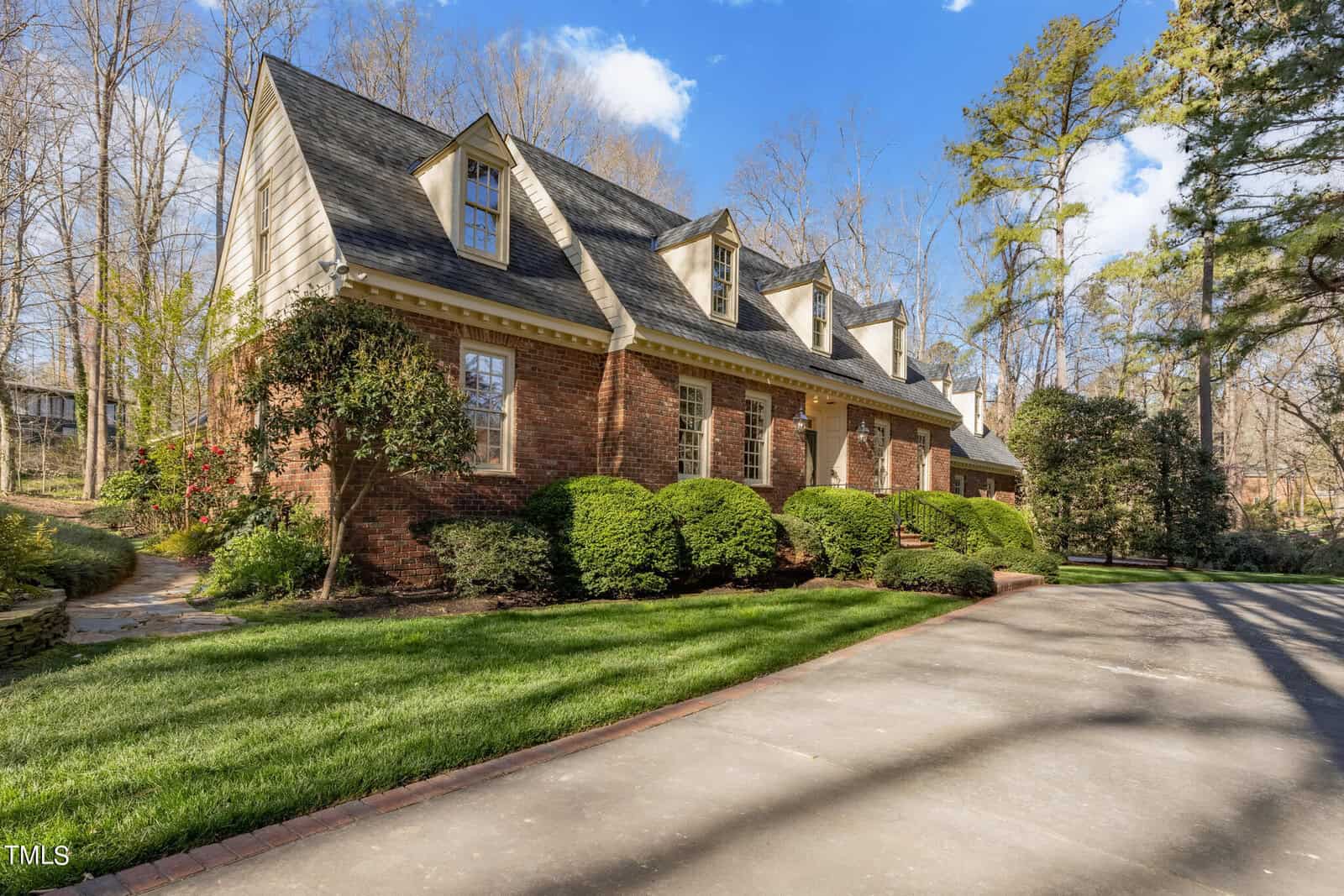 A charming two-story brick house with multiple gabled roofs, surrounded by lush green landscaping and mature trees under a clear blue sky.