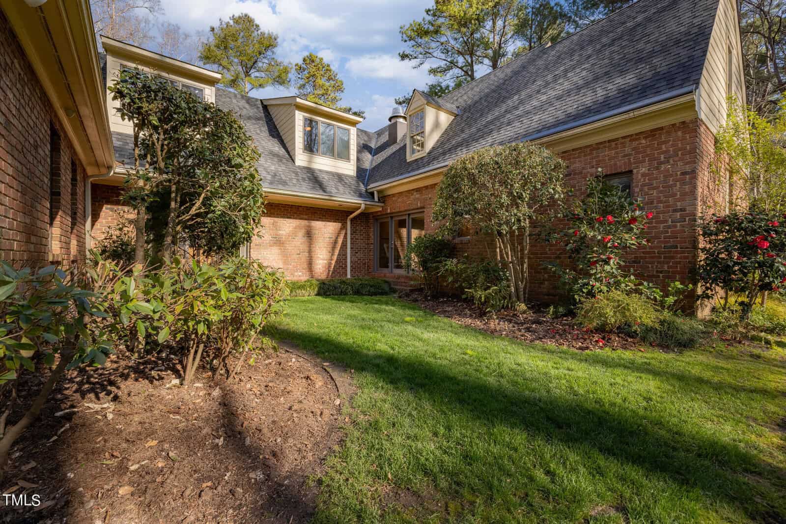 A large brick house with a sloping shingle roof surrounded by a landscaped garden with lush plants and a winding mulch path under a clear blue sky.