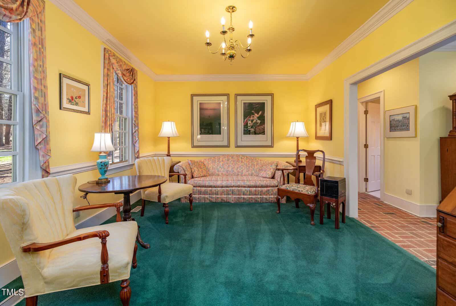 Brightly lit traditional sitting room with yellow walls, teal carpet, floral sofa, white armchairs, wooden tables, and large windows. decor includes lamps, paintings, and a chandelier.