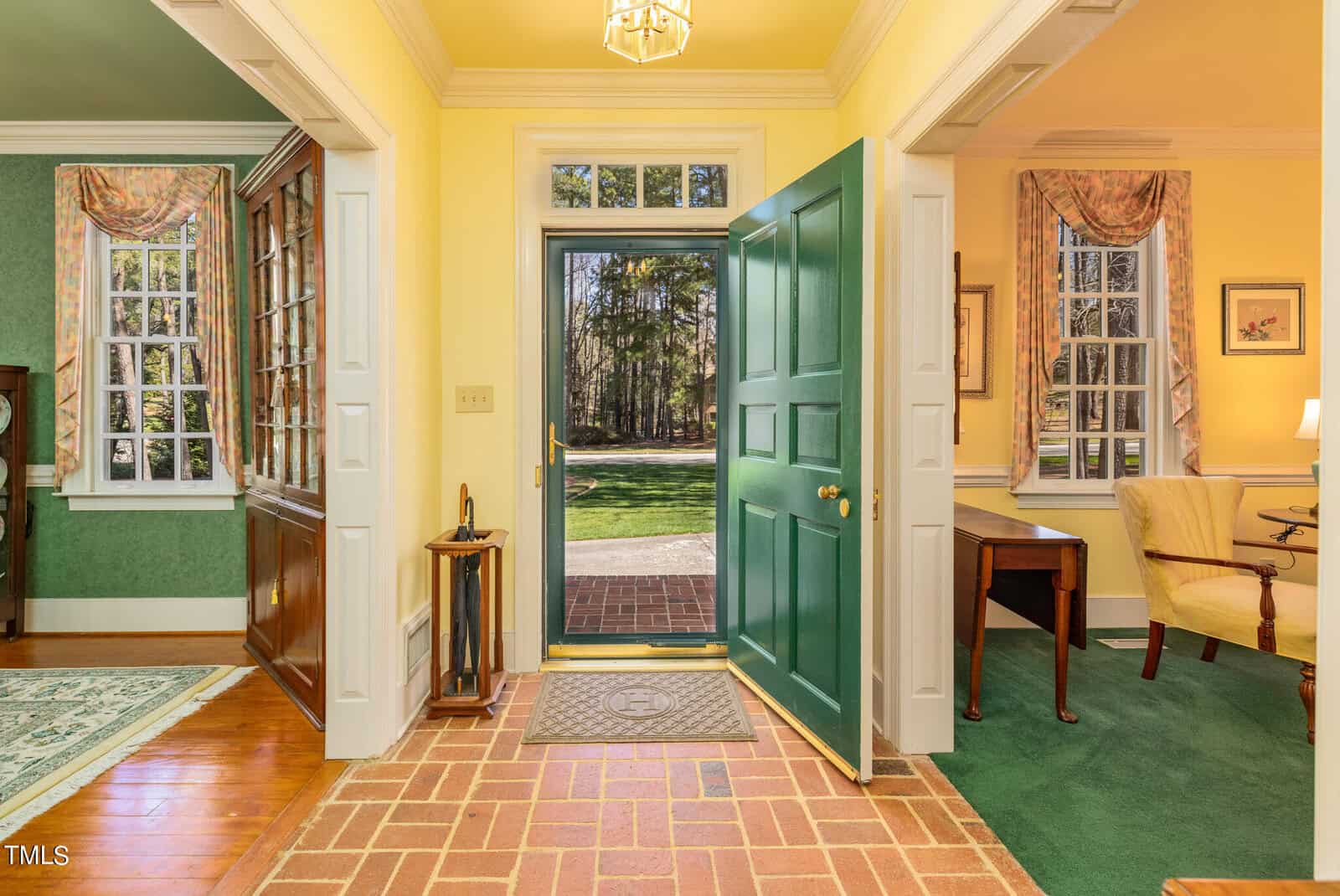 Elegant home interior showcasing an open green door leading to a lush garden, with a view of a desk and bright yellow walls, detailed with white and green moldings and window curtains.