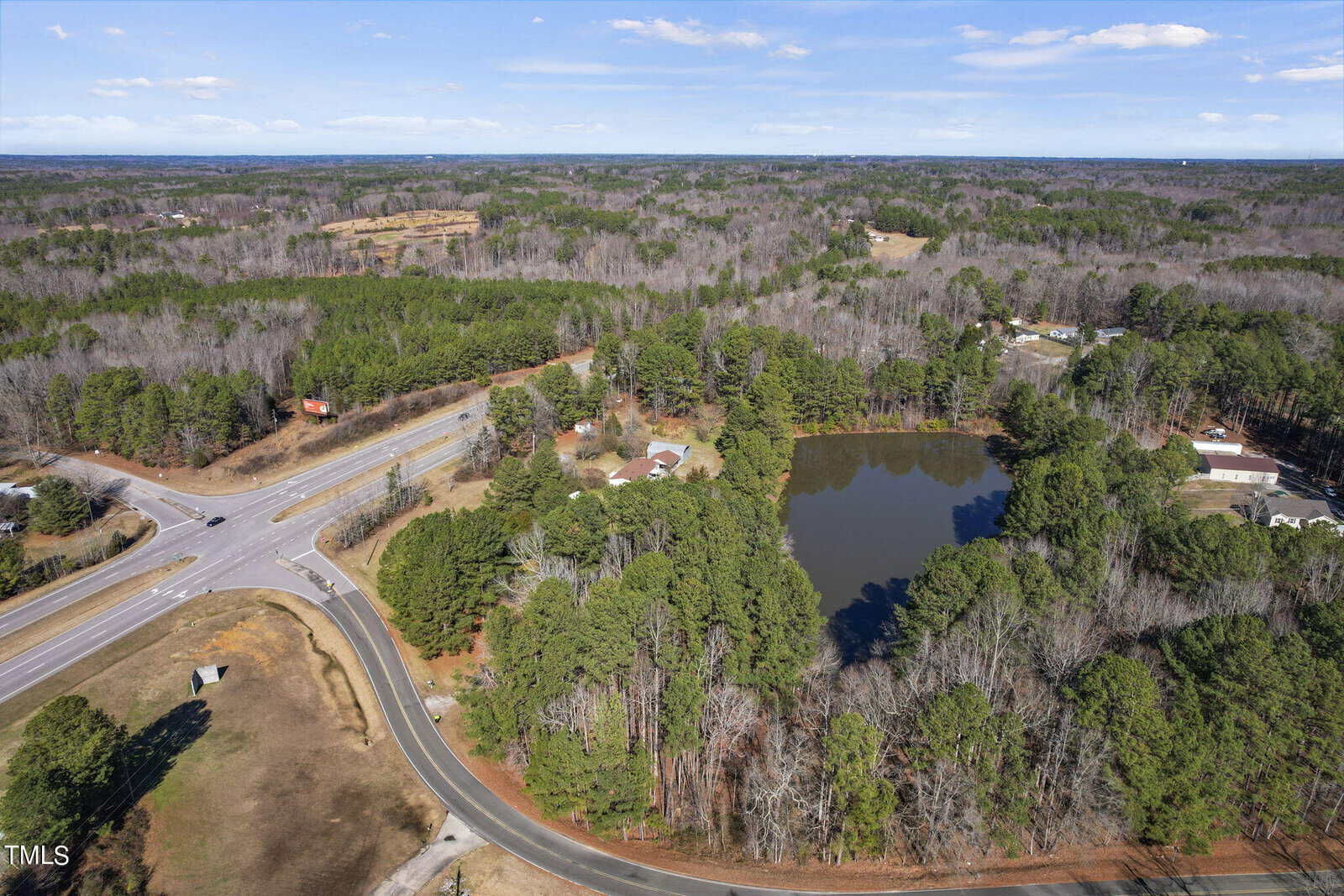 Aerial view of a wooded area with a road intersection, featuring a pond surrounded by trees, and residential properties scattered around under a clear blue sky.