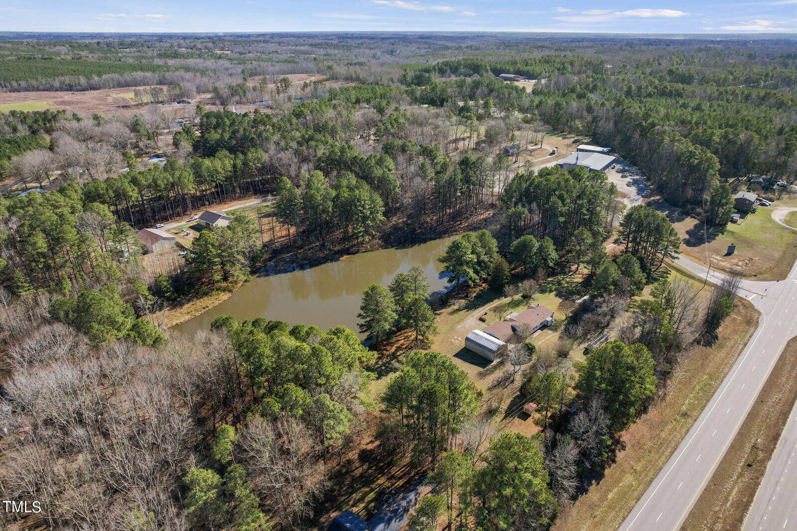 Aerial view of a small lake bordered by trees and roads with several buildings located around it in a rural setting, showcasing patches of forests and clear land stretching into the distance near 51 S Lake Lodge