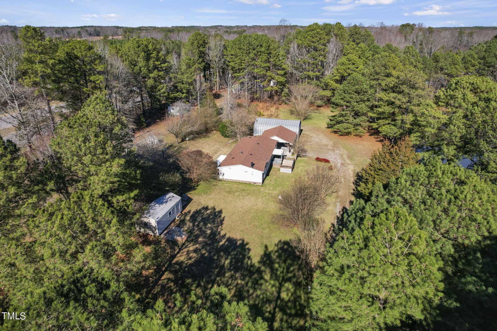 Aerial view of a house with a brown roof surrounded by dense green trees, featuring a large open yard and an adjacent white trailer.