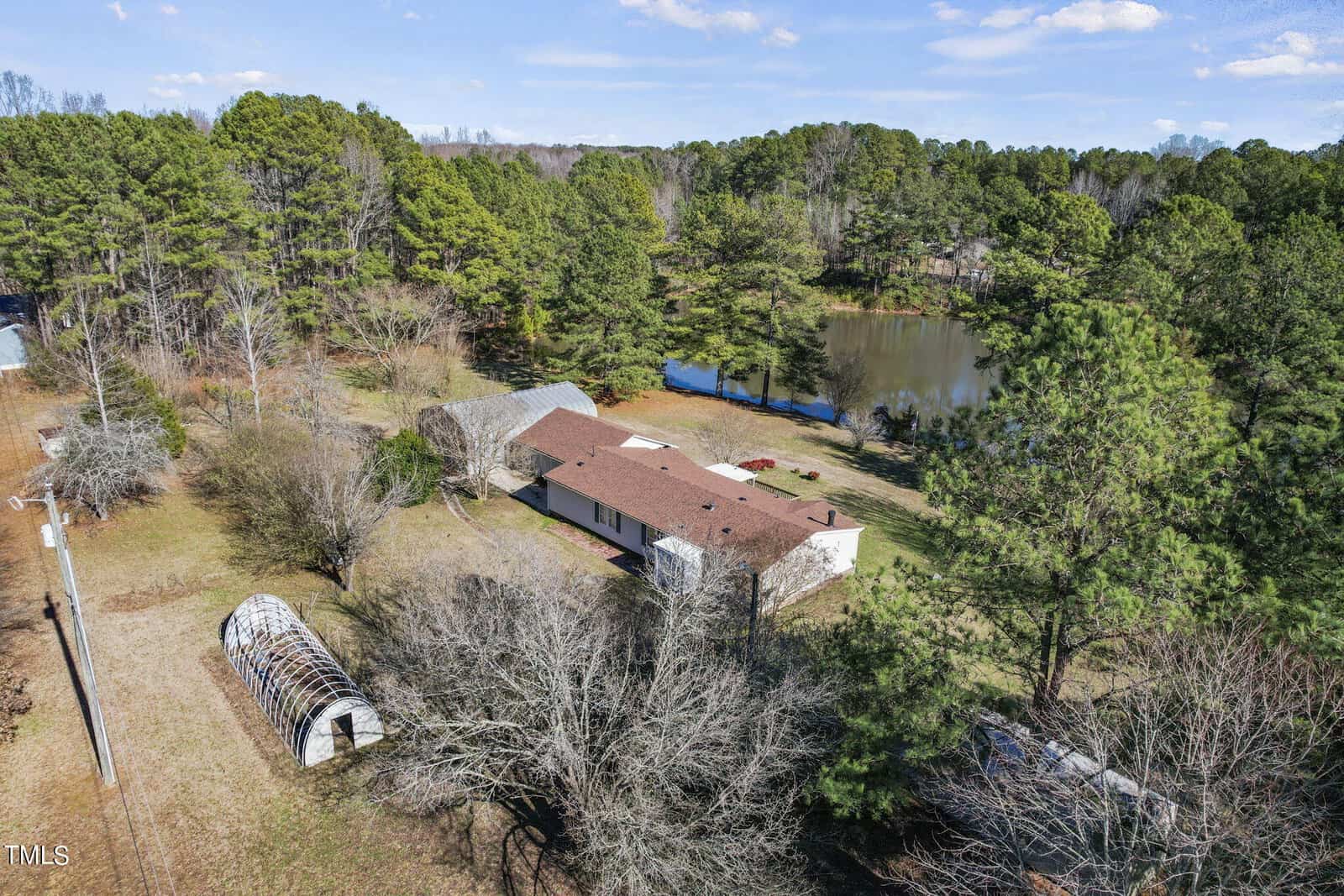 Aerial view of a rural property featuring a single-story house, a small pond, and dense surrounding trees. a curved, metallic structure stands near the residence.
