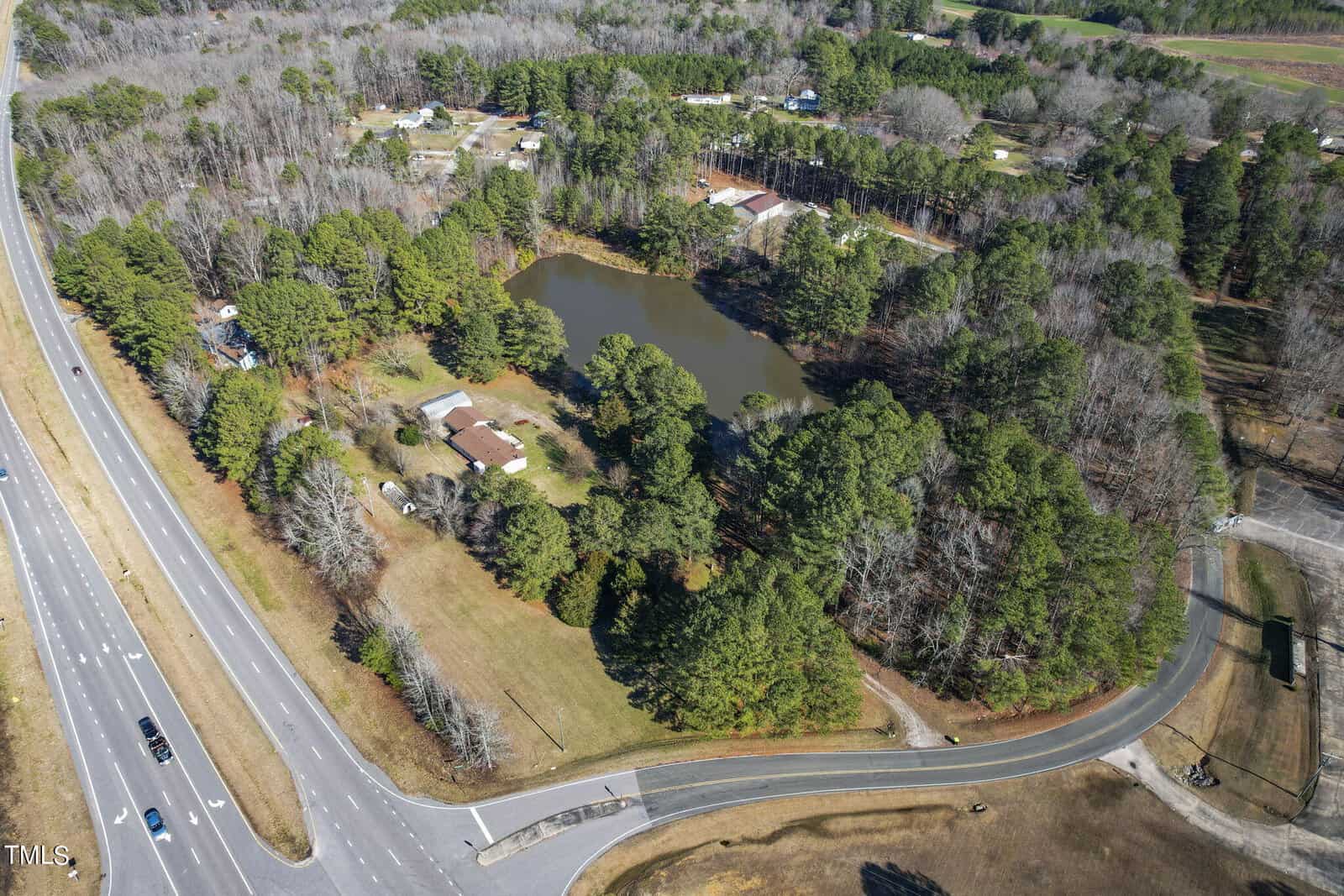 Aerial view of a serene pond surrounded by trees, with adjacent roads and residential structures nestled in a rural landscape.