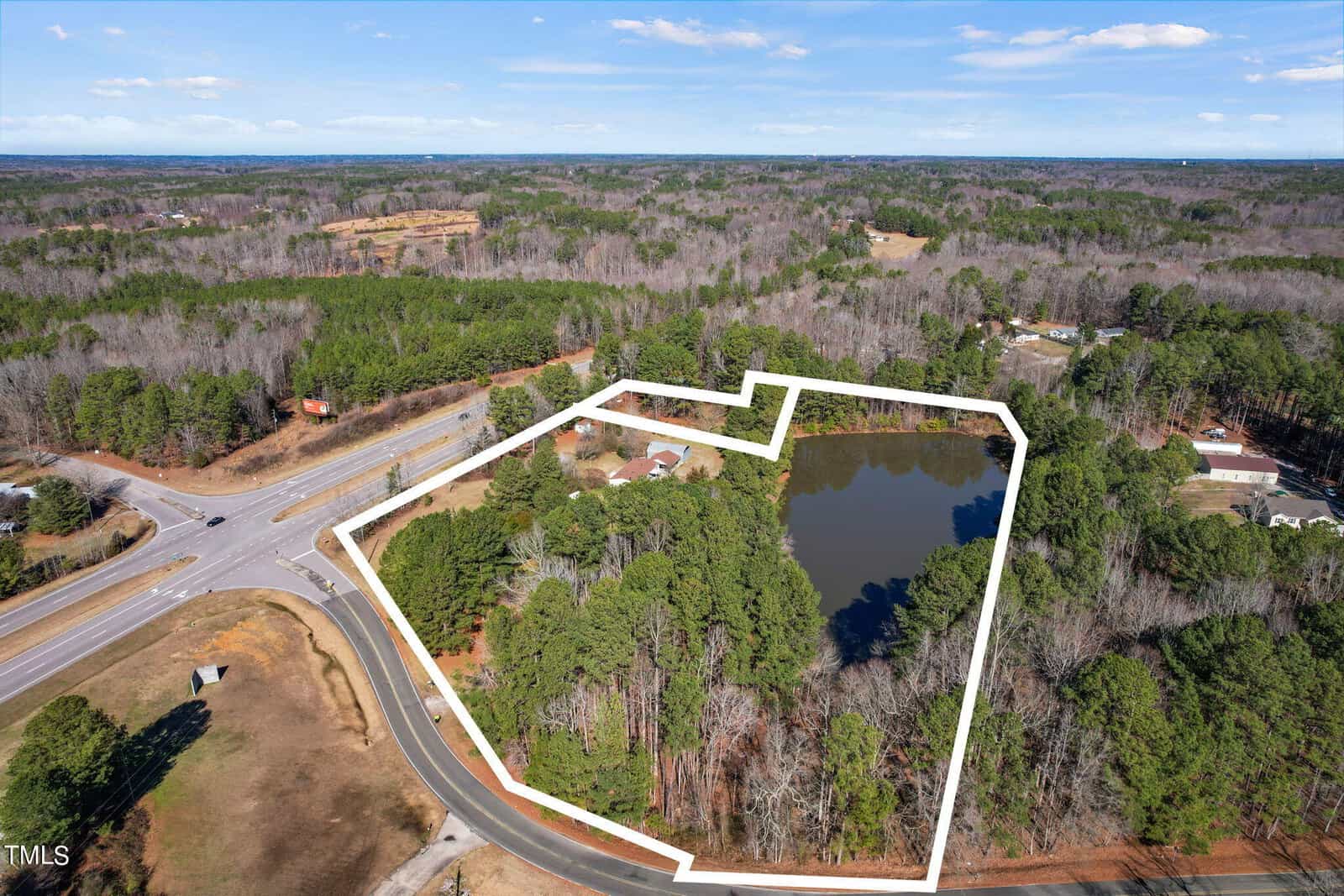 Aerial view of a property outlined in white, featuring a pond surrounded by trees near a road intersection, with distant views of a suburban area.