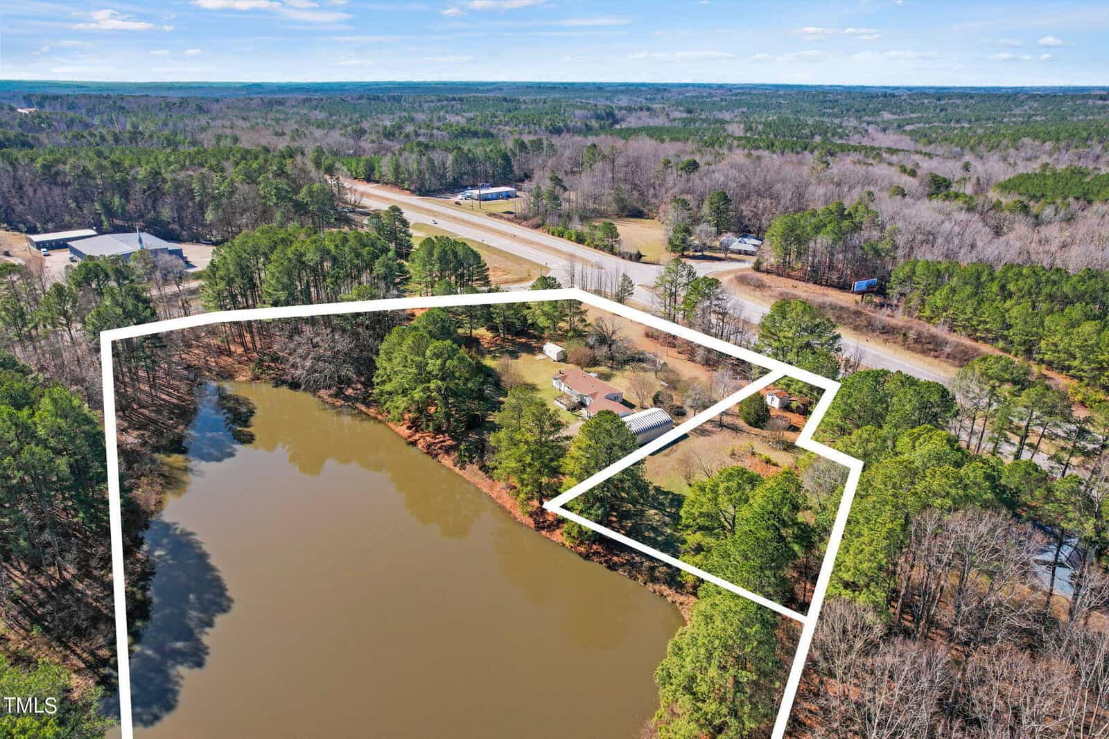Aerial view of a property outlined in white, featuring a large pond, several buildings, and surrounded by trees near a highway.