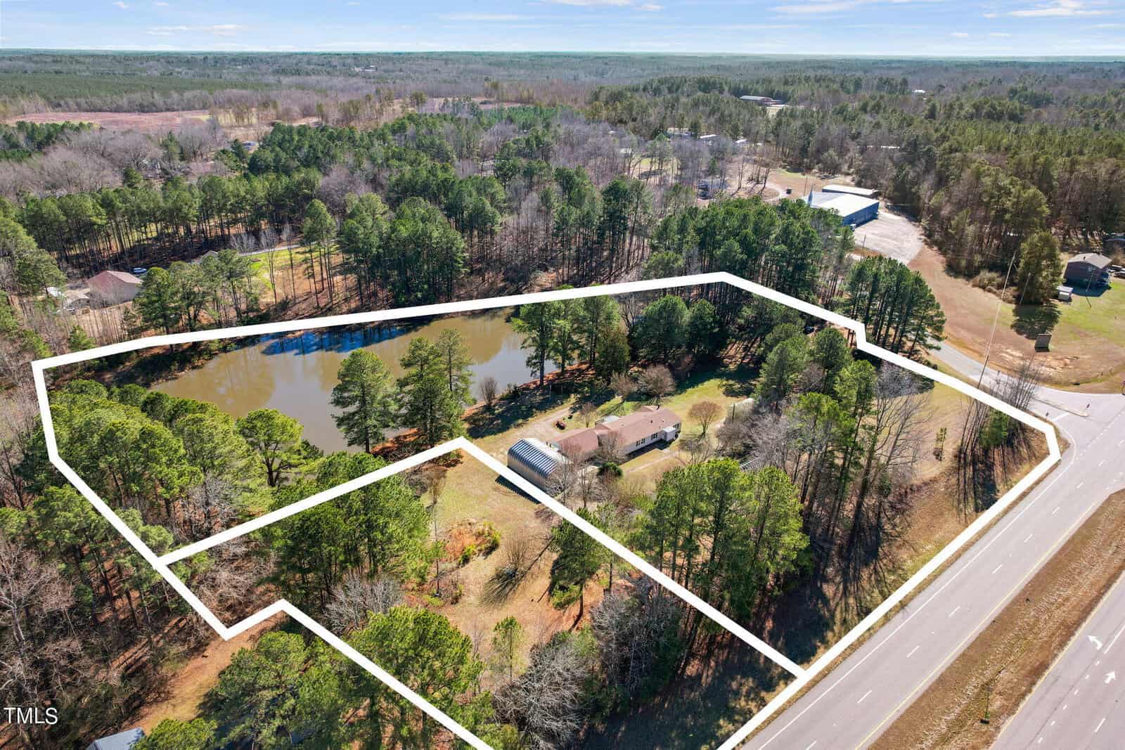 Aerial view of a property outlined by a white line, featuring a pond, a house, and a large shed surrounded by trees near a roadway.