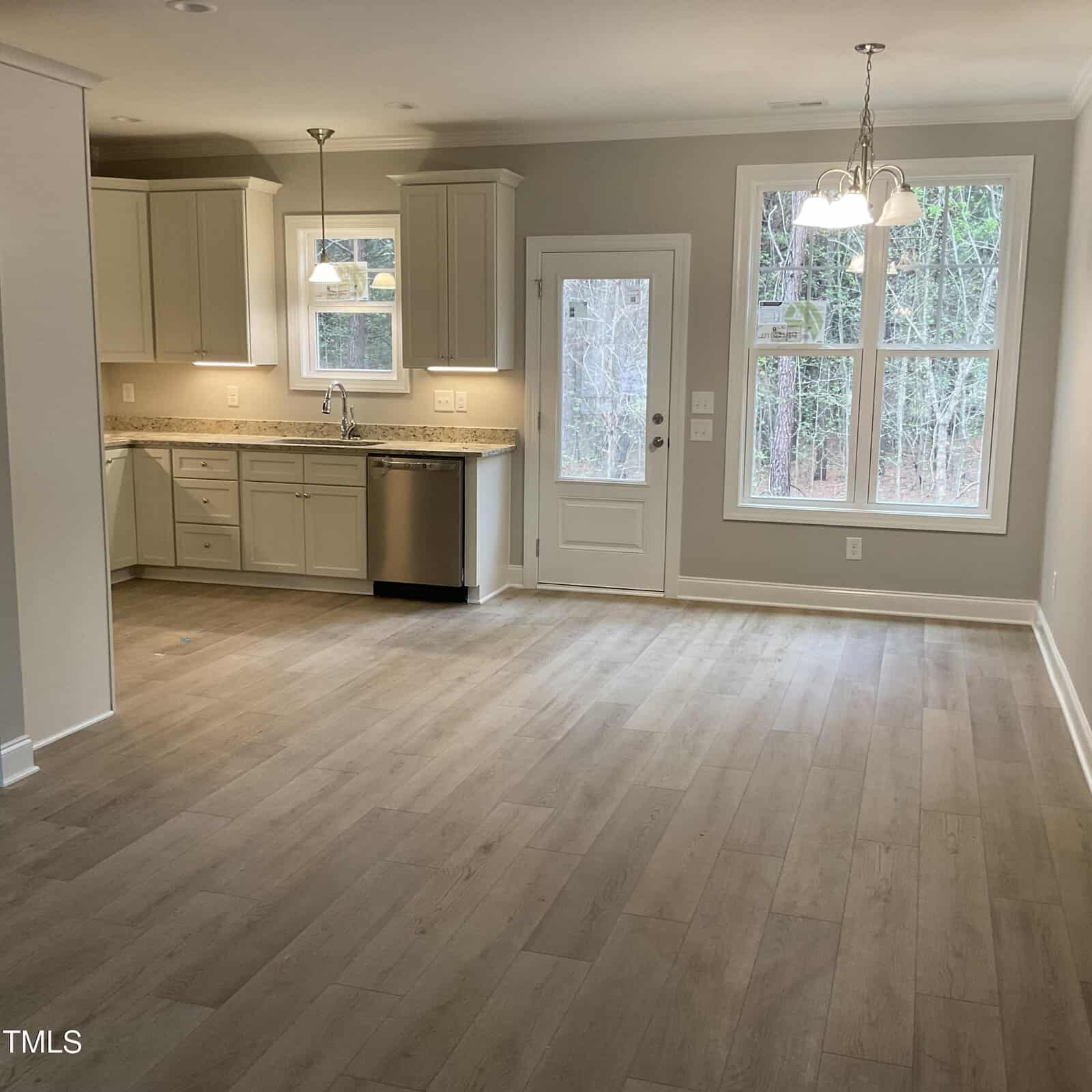 A modern kitchen with light wood flooring, white cabinets, granite countertops, and a stainless steel dishwasher. Two pendant lights hang above the counter, and a window and glass door offer a view of trees outside.