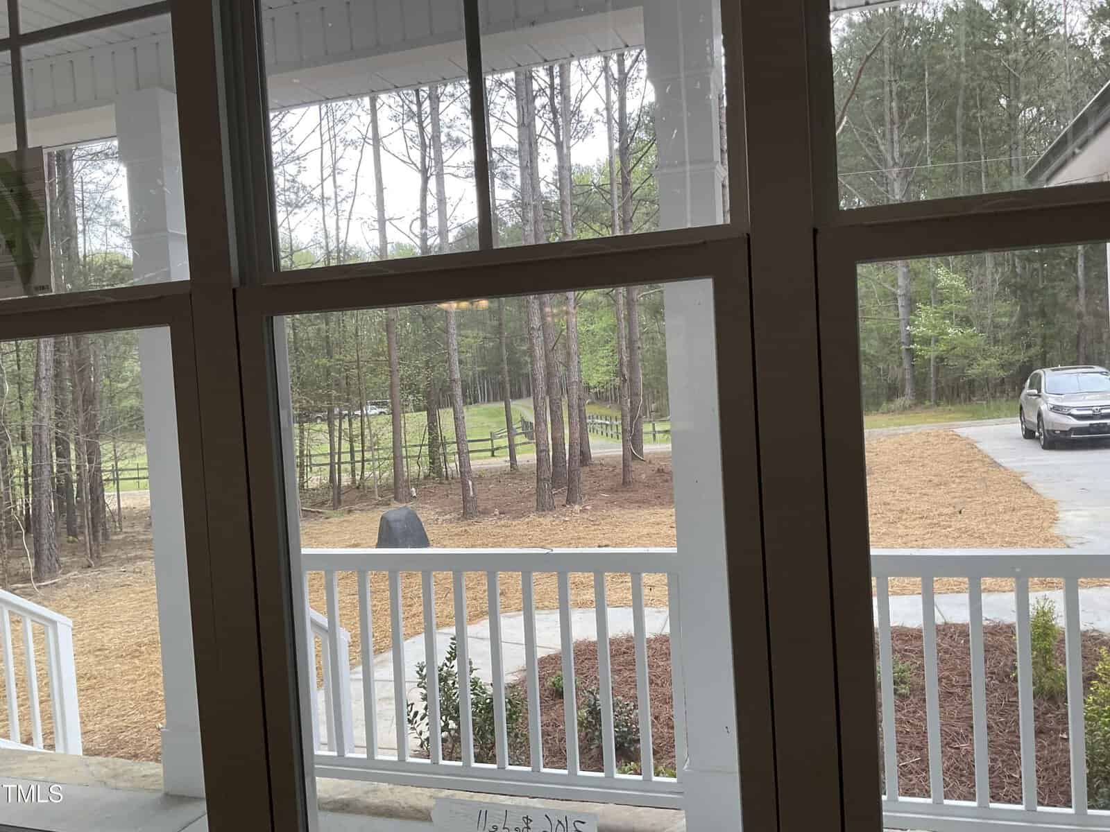 View through a window showing a front porch with white railings, a driveway, and a car parked on the side. Beyond the porch, there are tall trees and a grassy area, creating a serene, wooded scene.