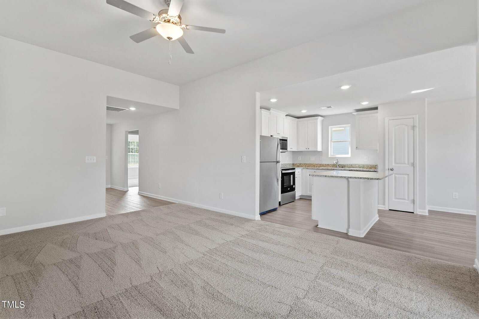 Spacious, unfurnished living room with light-colored carpet, ceiling fan, and an open design leading to a modern kitchen with white cabinetry, stainless steel appliances, and a breakfast bar. Adjacent are a corridor and a doorway leading to another room.