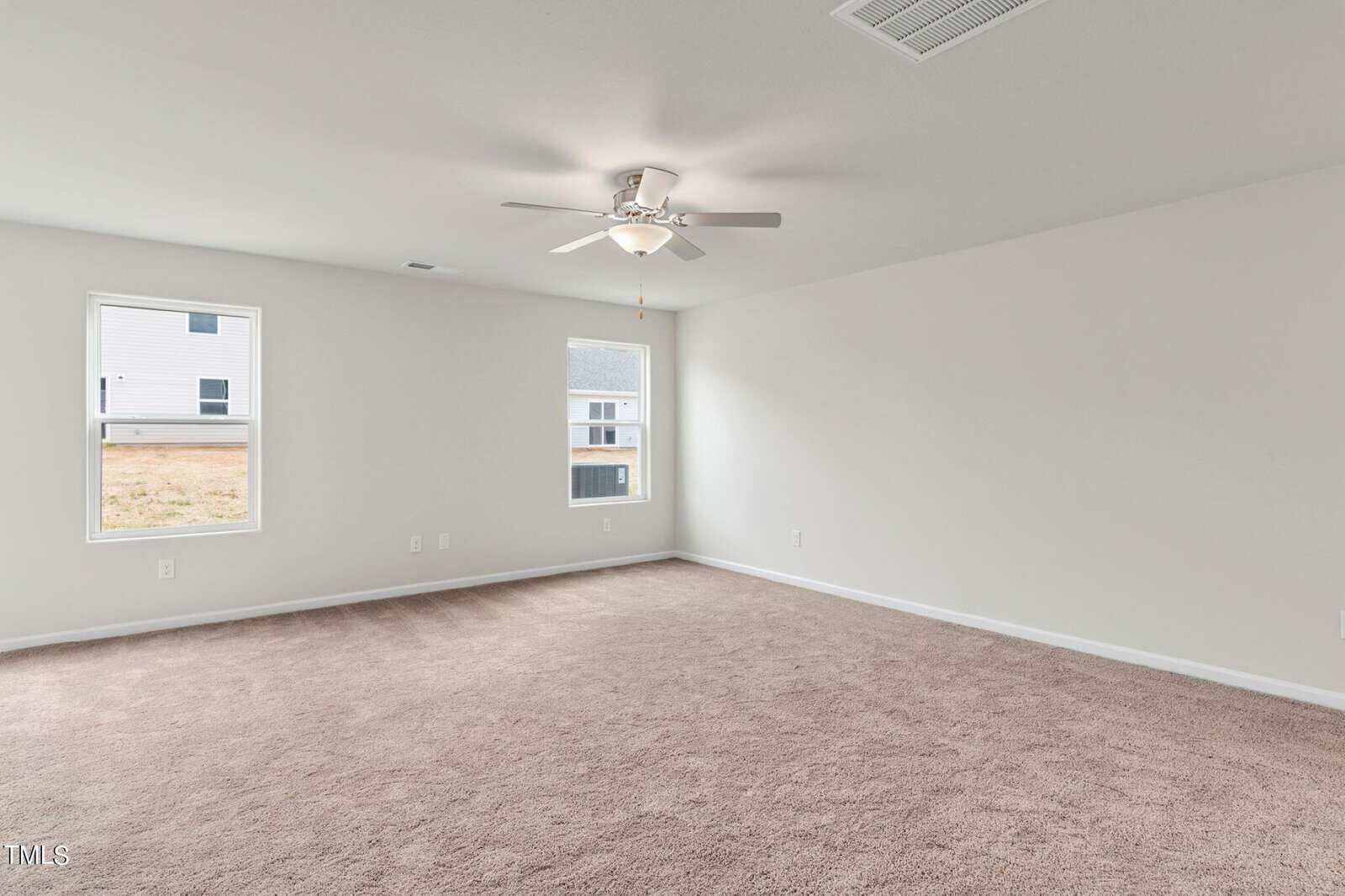 A spacious, empty room with beige carpet, white walls, and a ceiling fan. Two windows let in natural light, with views of a neighboring house and a grassy area outside.