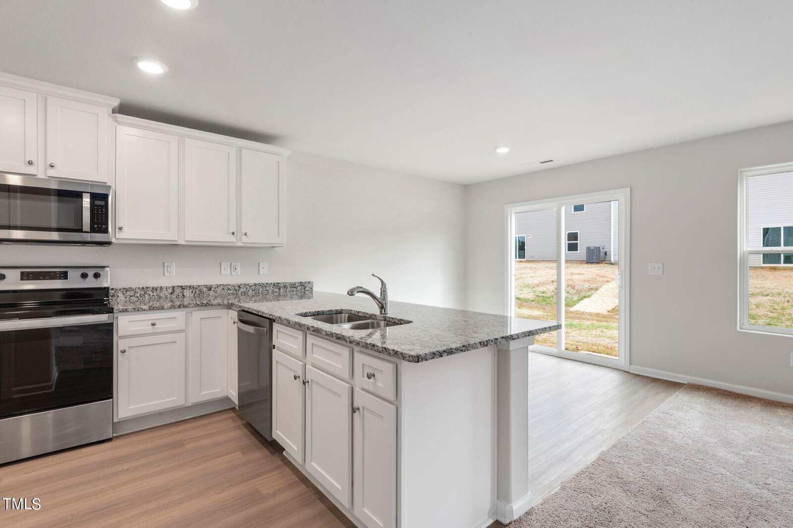 A modern kitchen with white cabinets, stainless steel appliances, and a granite countertop island with a sink. The space opens to a bright living area with large sliding glass doors leading to a backyard. Hardwood and carpeted flooring are visible.