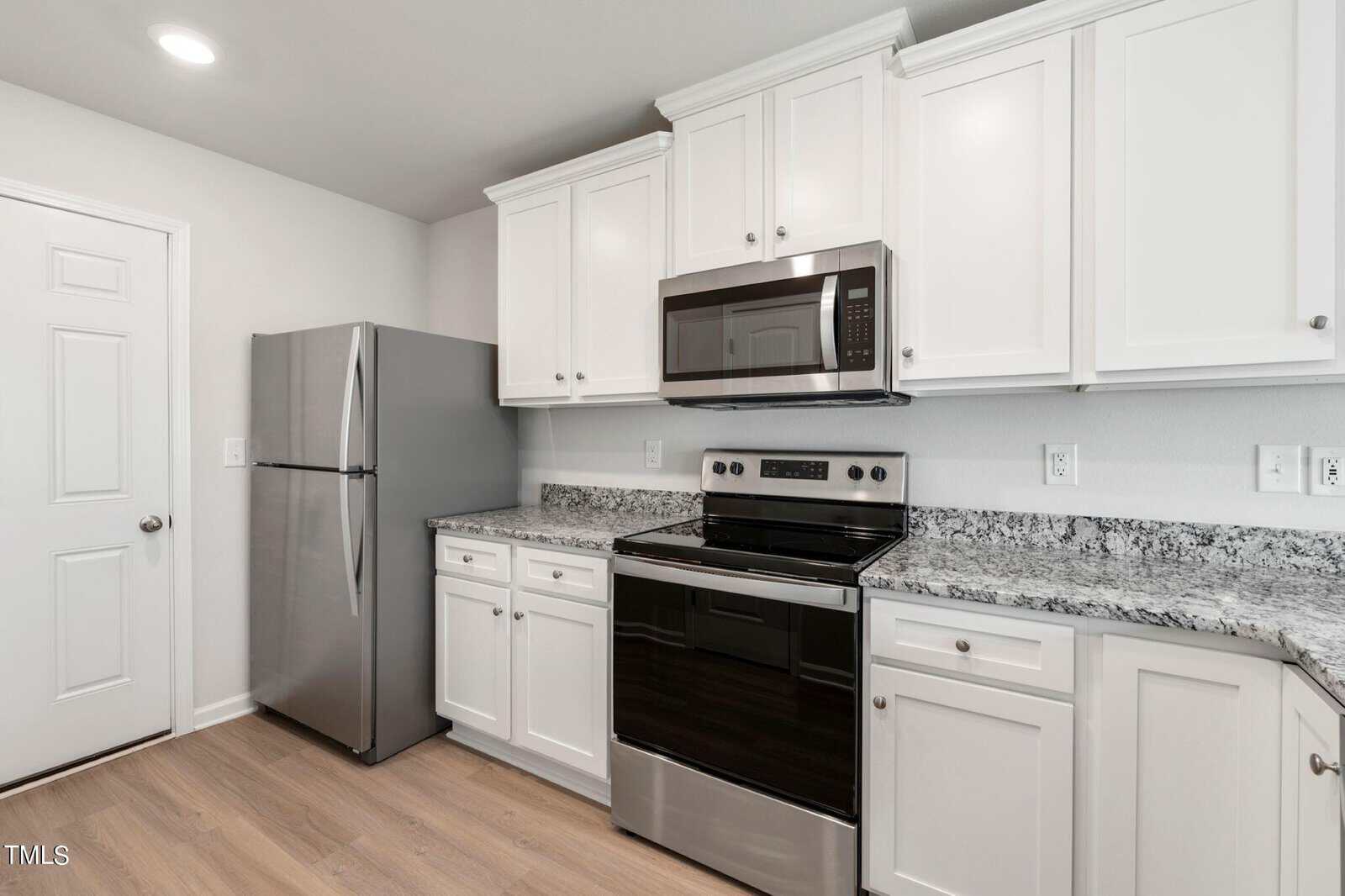 A modern kitchen featuring white cabinets, a stainless steel refrigerator, and an electric stove with an overhead microwave. The countertop is gray granite, and the flooring is light wood. A closed white door is on the left.
