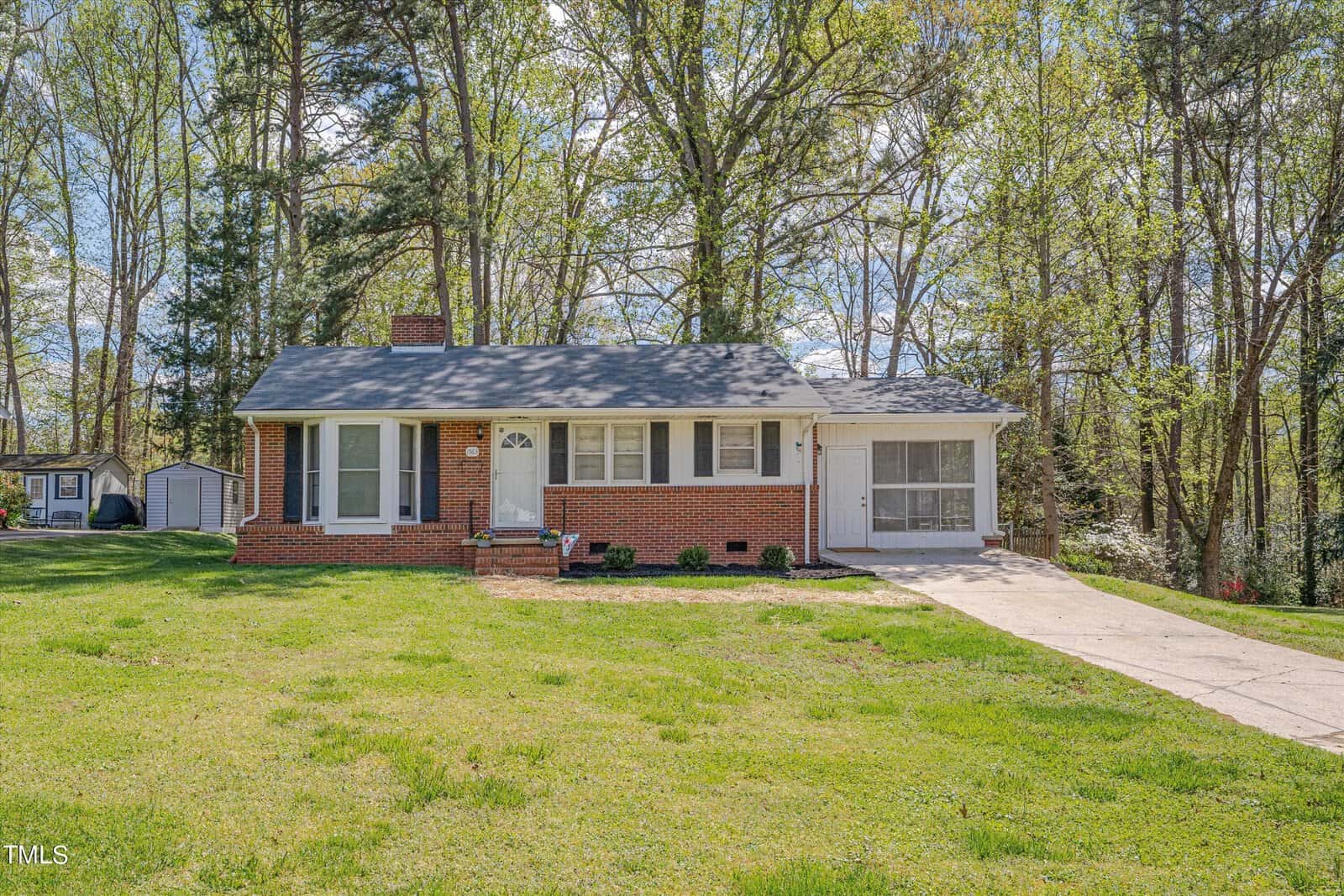 A one-story brick house with white trim, located at 1563 Oakdale Circle in Henderson, NC, features a gabled roof and an attached garage. The home is surrounded by a lush,