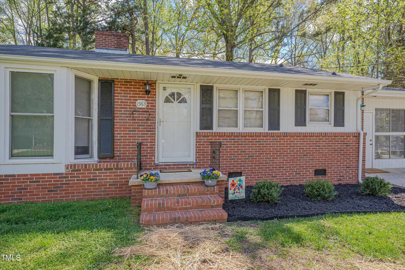 A single-story brick house with white siding under large windows and a central door, surrounded by trees, featuring a small front porch with a flowerpot and a yard with fresh mulch.