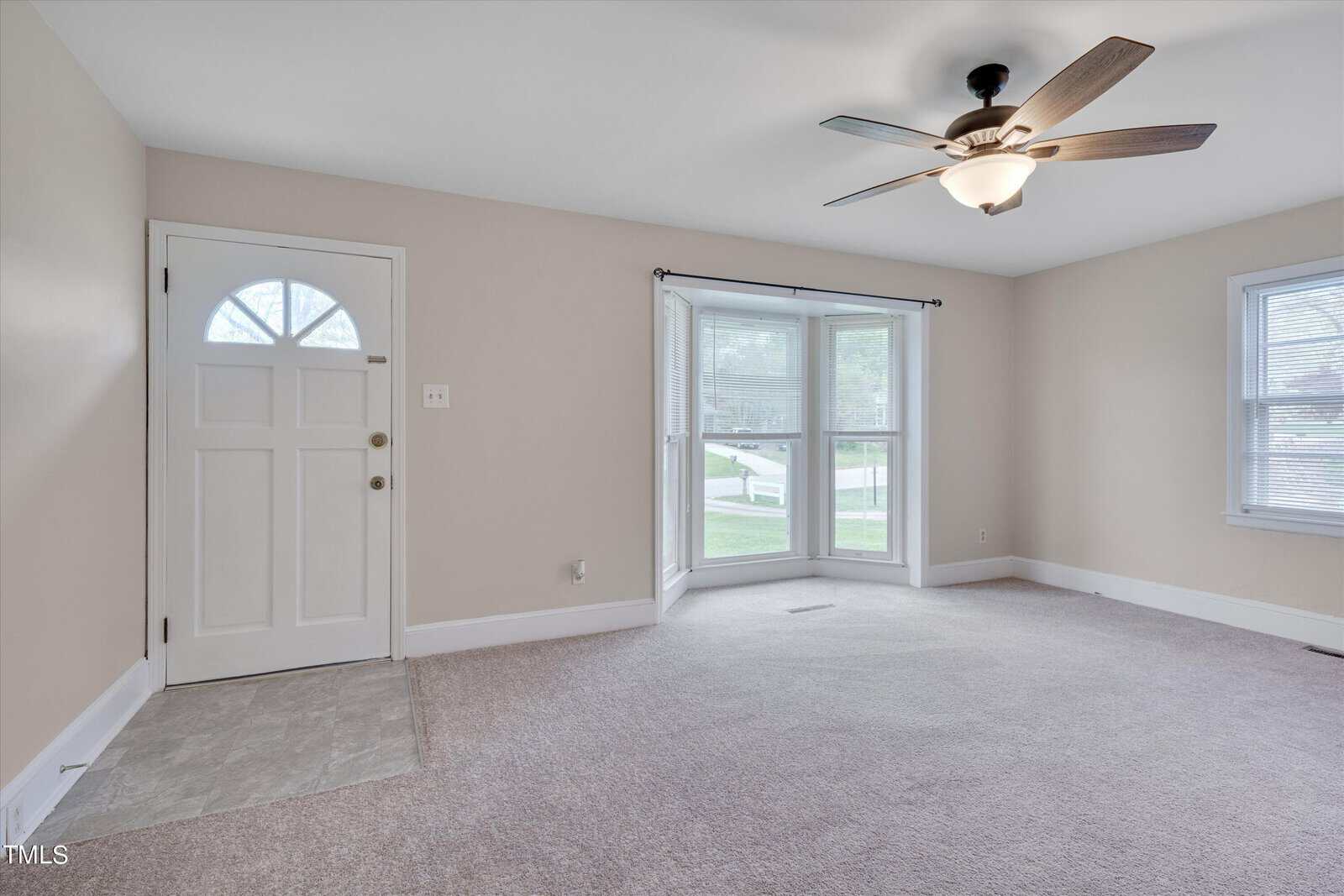 A clean and empty room featuring beige walls and carpet, with a ceiling fan overhead. it has a white door on one side and a large sliding glass door opening to the outside on the other.