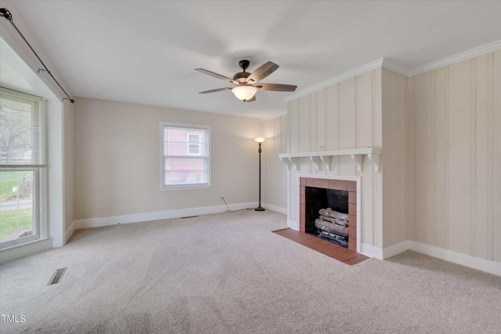 Bright, spacious living room with beige carpet, white walls with partial wood paneling, a fireplace, a ceiling fan, and two windows with street view.