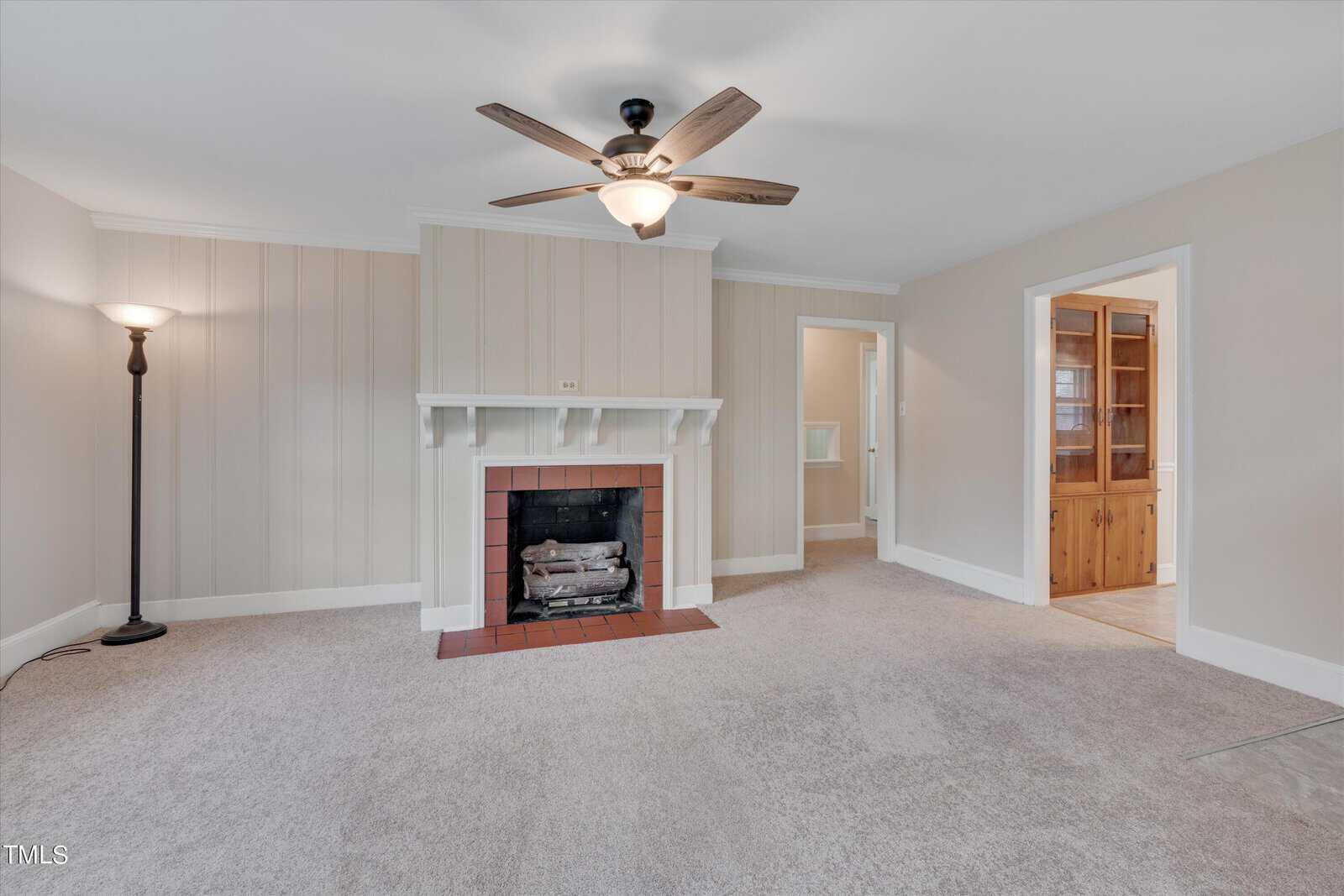 A cozy living room featuring a white fireplace with a brick hearth, beige carpeting, white wainscoting, and a ceiling fan. two doorways lead to adjacent rooms. a floor lamp stands on the left.