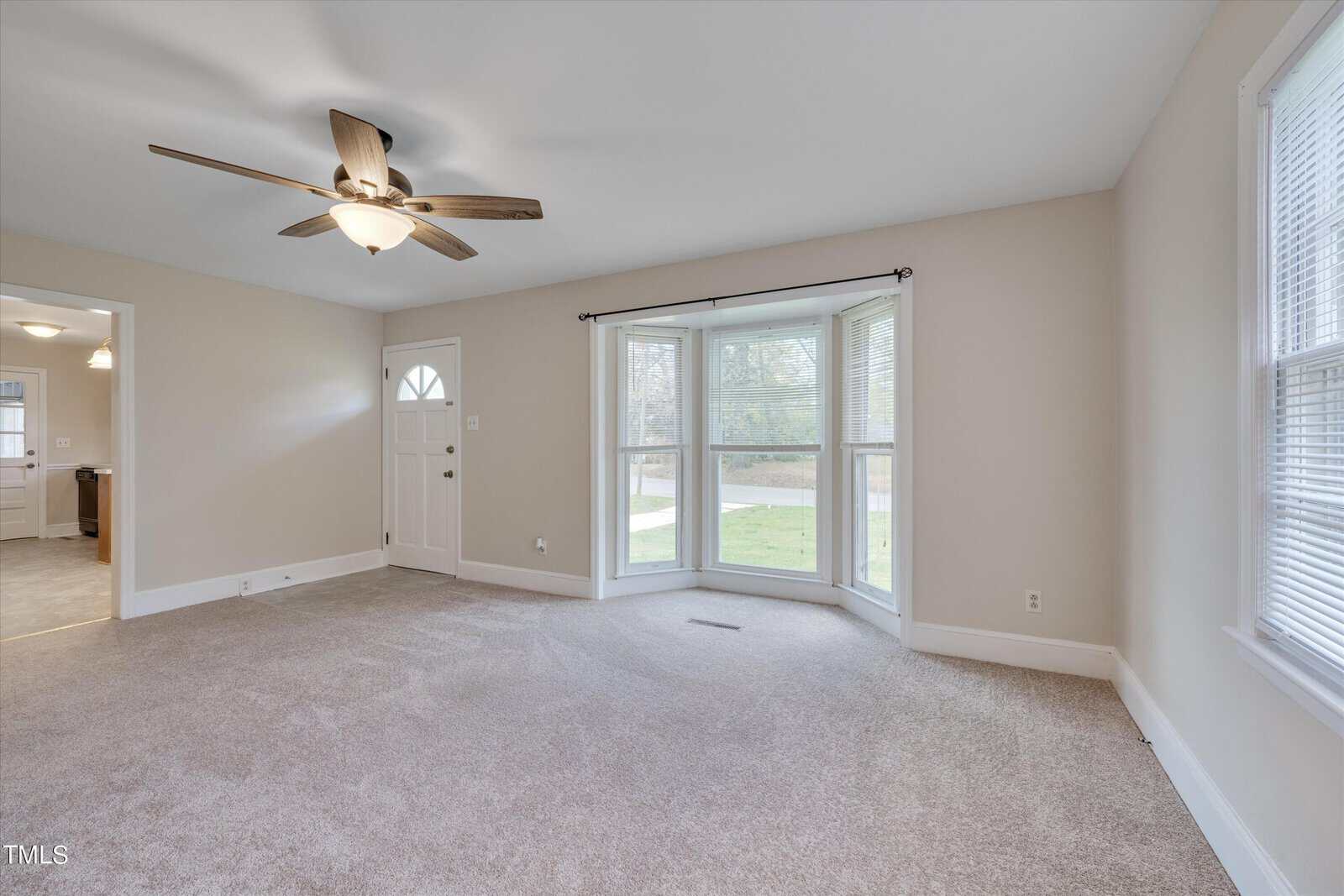 An empty, brightly lit room with beige carpet and white walls. it features a ceiling fan, large windows, and a sliding glass door leading to the outside.