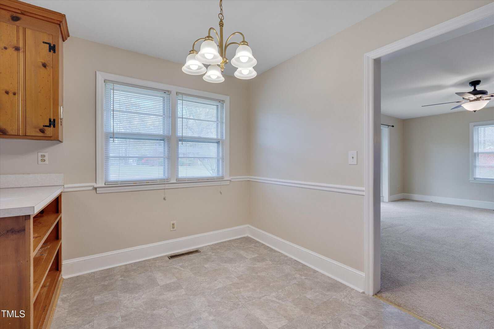 An empty room featuring light gray walls with white trim, beige tile flooring, and a corner wooden cabinet. there are two windows with blinds and a chandelier overhead.