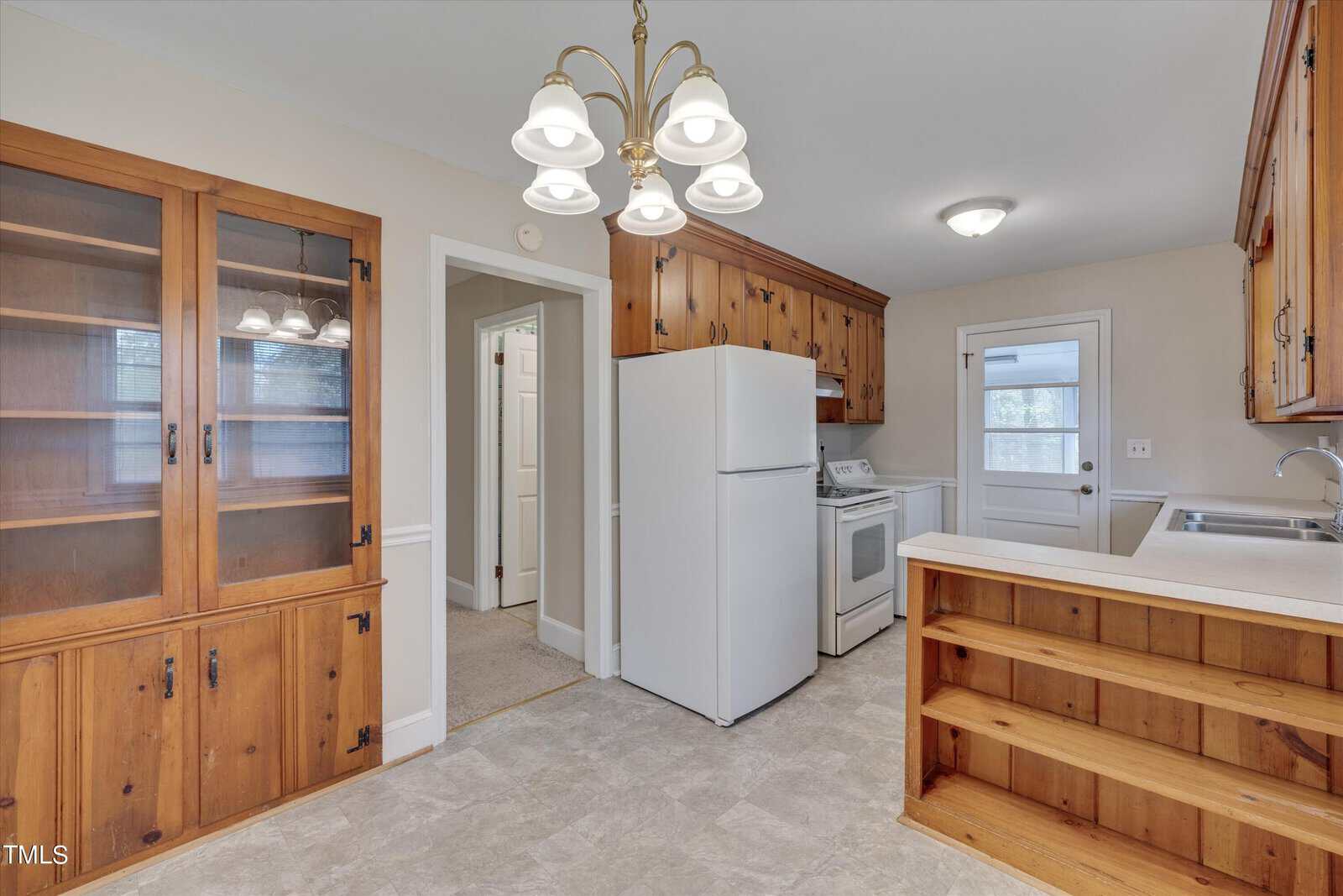 A spacious kitchen with wooden cabinetry, a white refrigerator and stove, and multiple doorways leading to adjacent rooms. the room features a tiled floor and a wooden breakfast bar.
