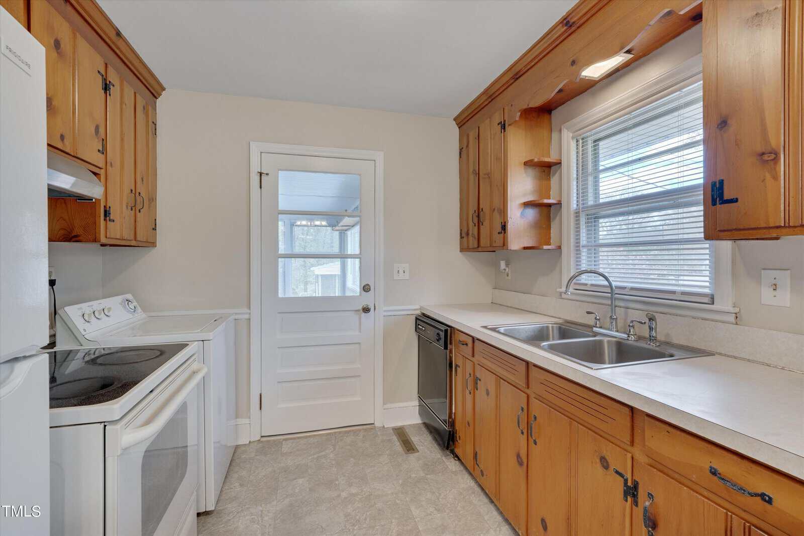 A small, well-lit kitchen with rustic wooden cabinetry, white walls, and gray tile flooring. features include a double sink beneath a window, a white stove, and a dishwasher.