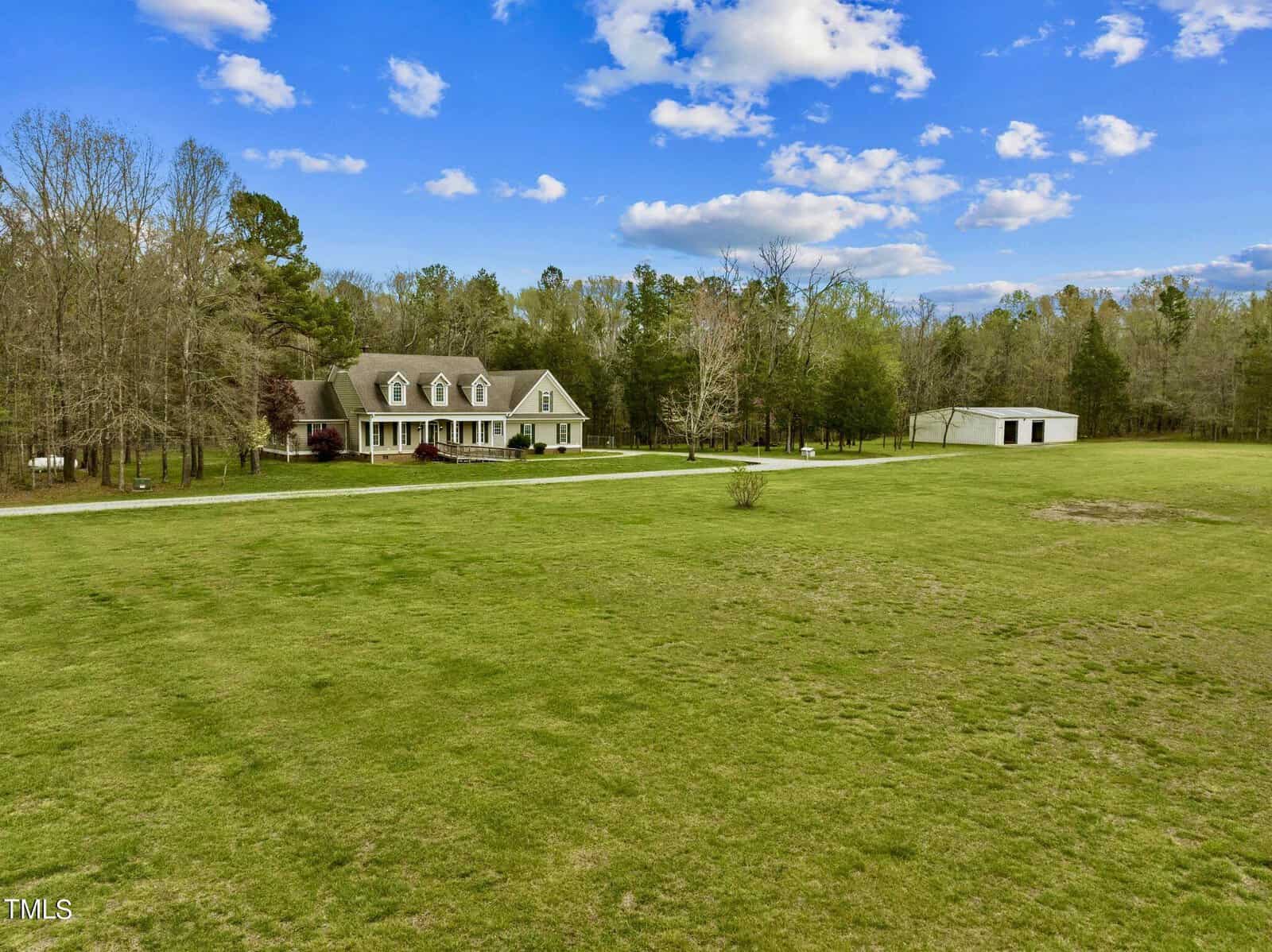 A large house with a gray roof sits amidst a vast green lawn under a blue sky with clouds. Trees surround the property, and there's a white barn-like structure to the side. A long driveway leads to the house.