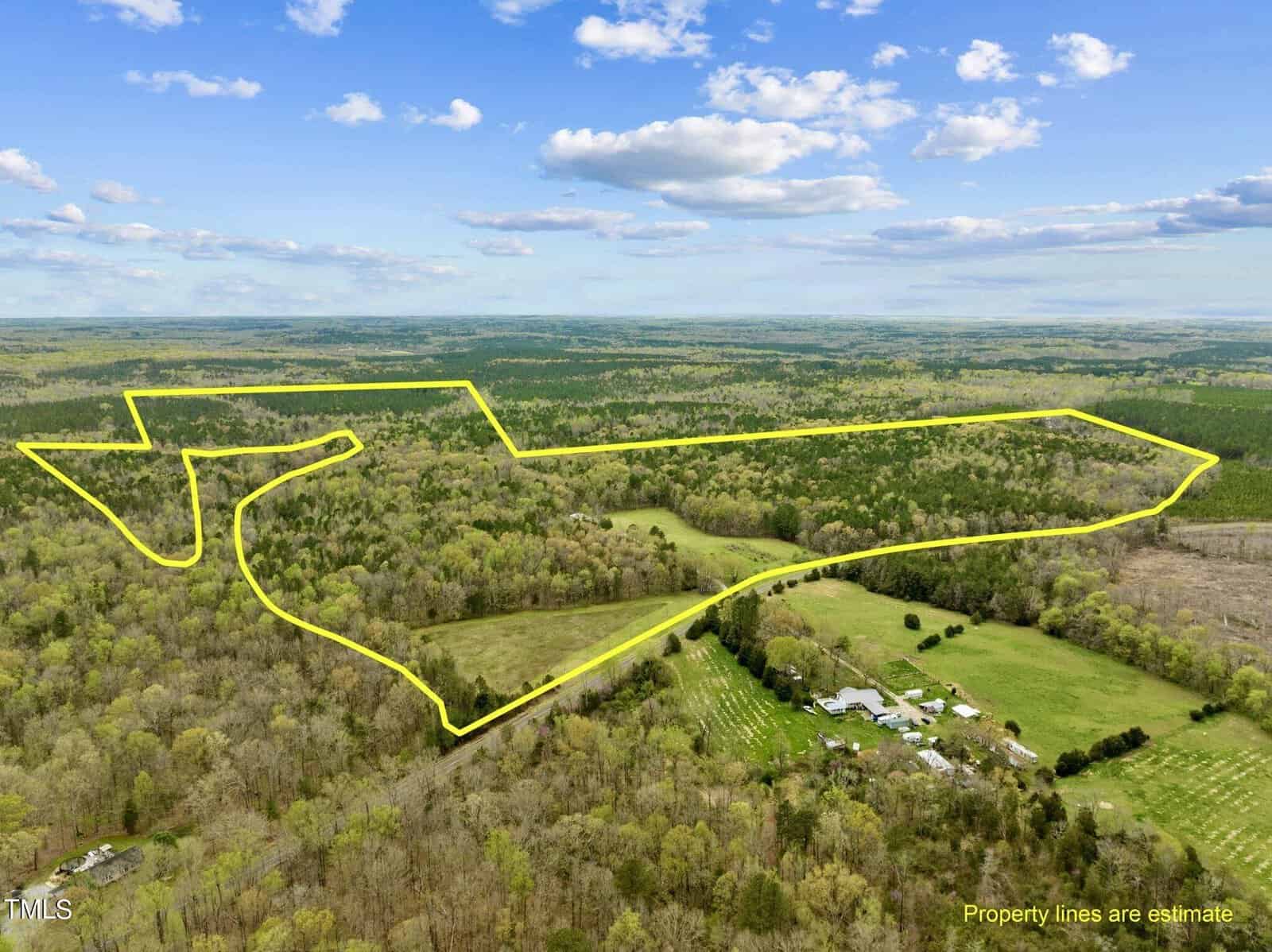 Aerial view of a lush, green landscape with dense forests and open fields. A yellow outline marks a large, irregularly shaped plot of land. A cluster of buildings is visible in the lower part of the image under a partly cloudy sky.