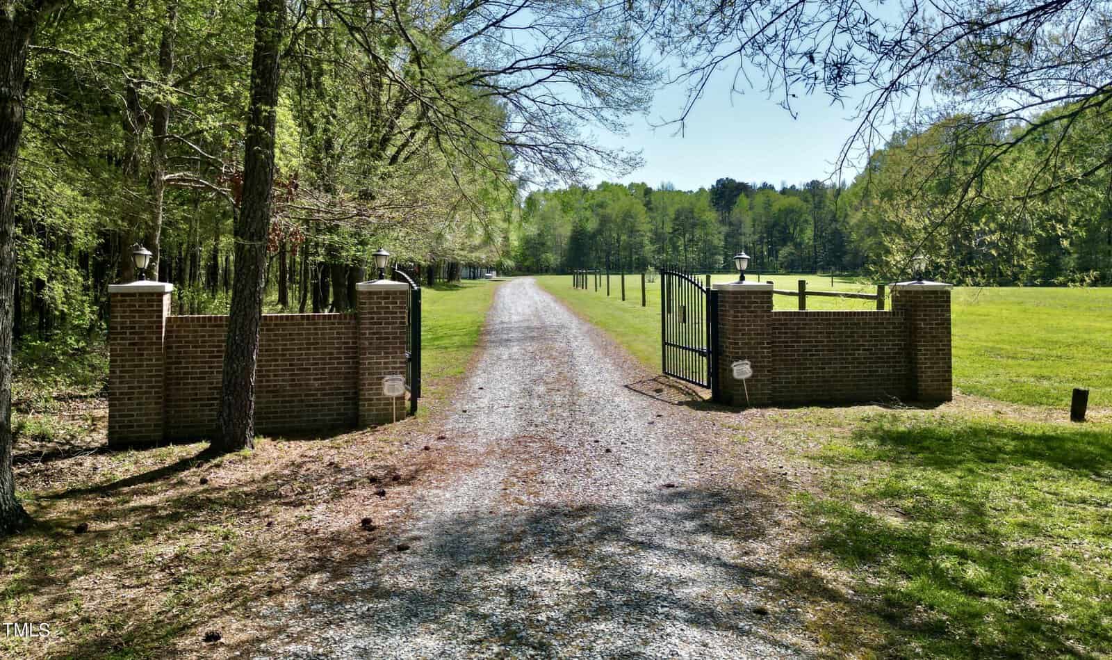 A gravel driveway leads through an open iron gate flanked by brick pillars, surrounded by green grass and tall trees. The path extends towards a wooded area under a clear blue sky.