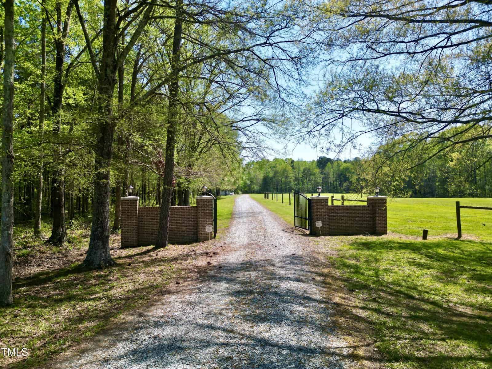 A gravel driveway leads to a distant building, flanked by open grassy fields and trees. The entrance is marked by brick pillars and surrounded by a lush green landscape under a blue sky.