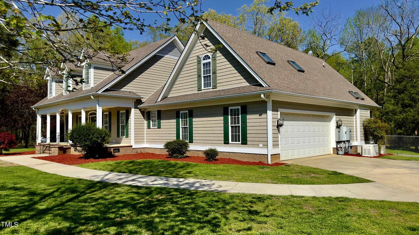 A two-story house with a steep gable roof and beige siding is surrounded by a well-manicured lawn. The house features multiple windows, a porch with white columns, and an attached two-car garage. Trees are visible in the background.