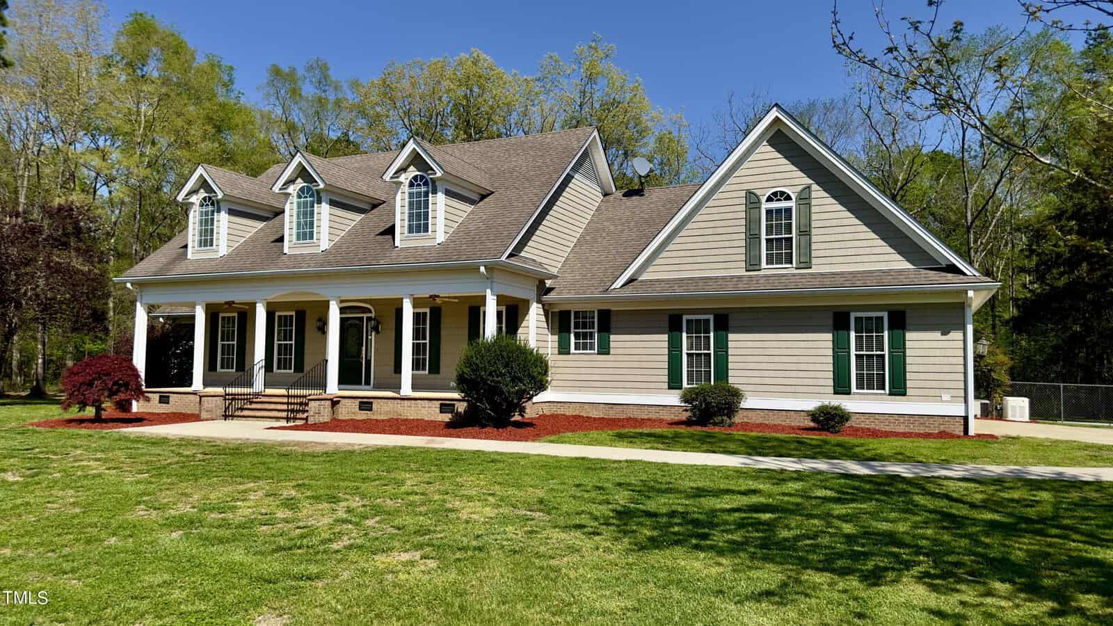 A two-story house with a steep roof, dormer windows, and a front porch. It's surrounded by a well-kept lawn, small bushes, and a few trees in the background. The sky is clear and blue.