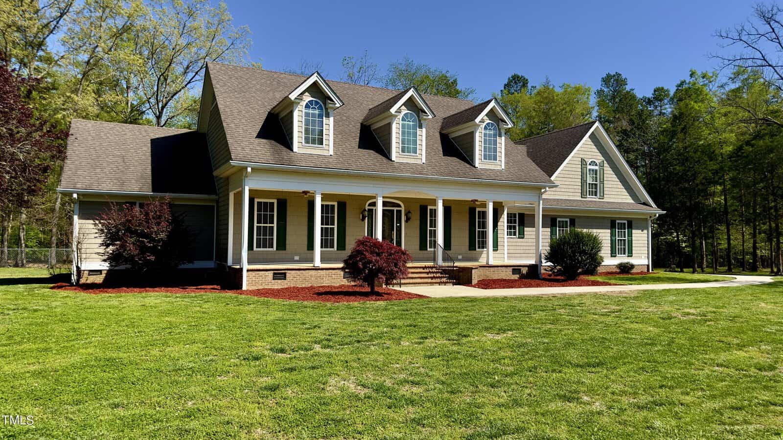 A spacious, traditional-style house with a well-maintained lawn, surrounded by trees. The home has a light exterior, dark roof, and several large windows, with a front porch supported by white columns.