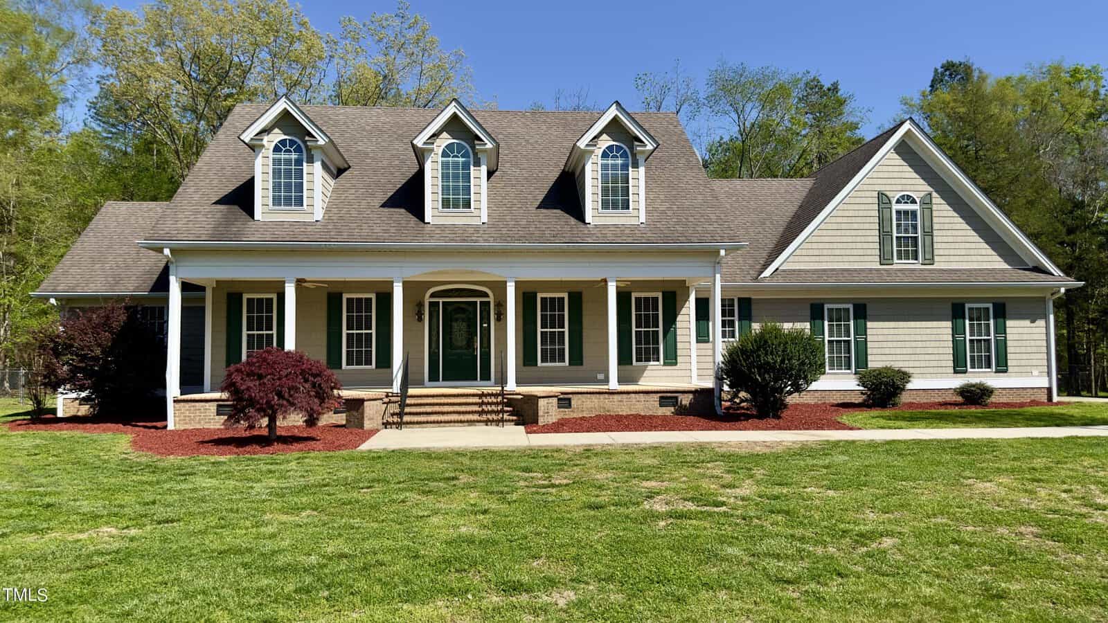 A two-story house with three dormer windows sits in a grassy yard. It features a large front porch with white columns and a central green door. Bushes and small trees are planted along the front, under a clear blue sky.