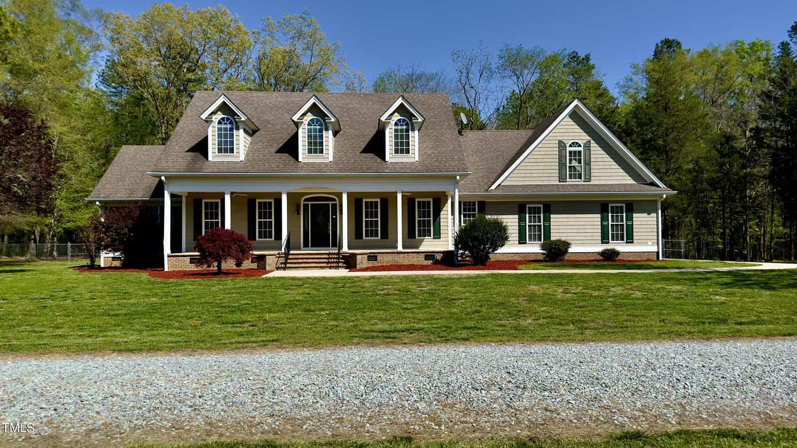 A two-story house with a gabled roof and three dormer windows. It has a covered front porch with white columns, surrounded by a manicured lawn and shrubs. A gravel driveway is in the foreground, and trees are in the background under a clear blue sky.