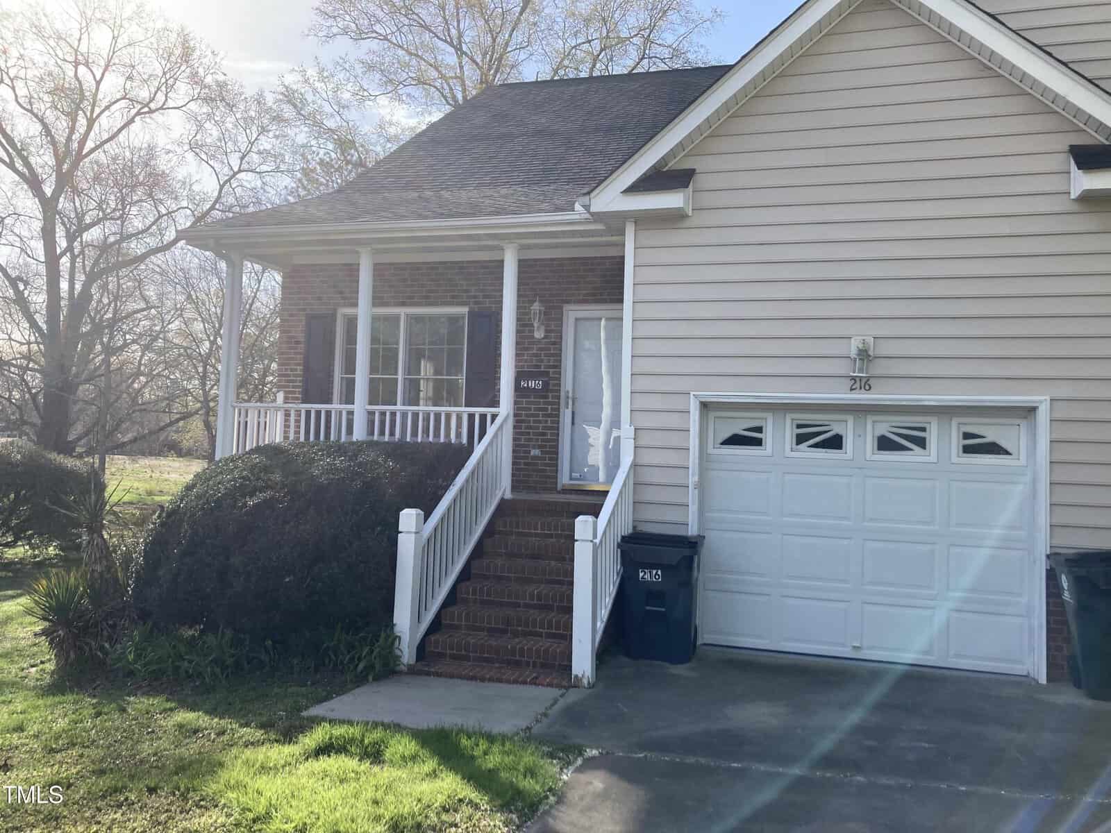 Located at 216 Clover Lane in the charming suburb of Oxford, this delightful house boasts a brick and siding exterior with a welcoming covered porch and white railing. A single-car garage is accessible via the driveway, all framed by lush trees and bushes.