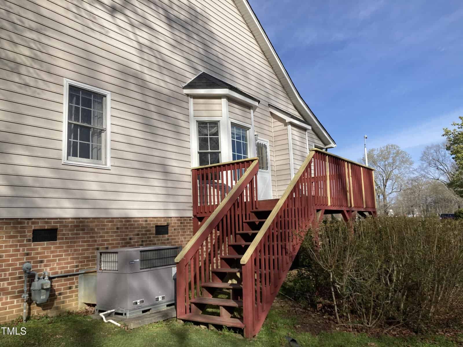 Side view of a house with beige siding and a brick foundation. A wooden staircase with red railing leads to a door. Bushes line the side, and an HVAC unit is visible. The sky is clear and blue.