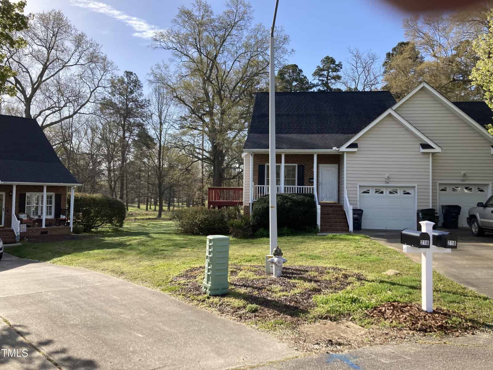 A suburban scene with two houses and a driveway. One house is brick with a porch, and the other is white with a double garage. Trees and a grassy area are visible in the background under a clear blue sky.